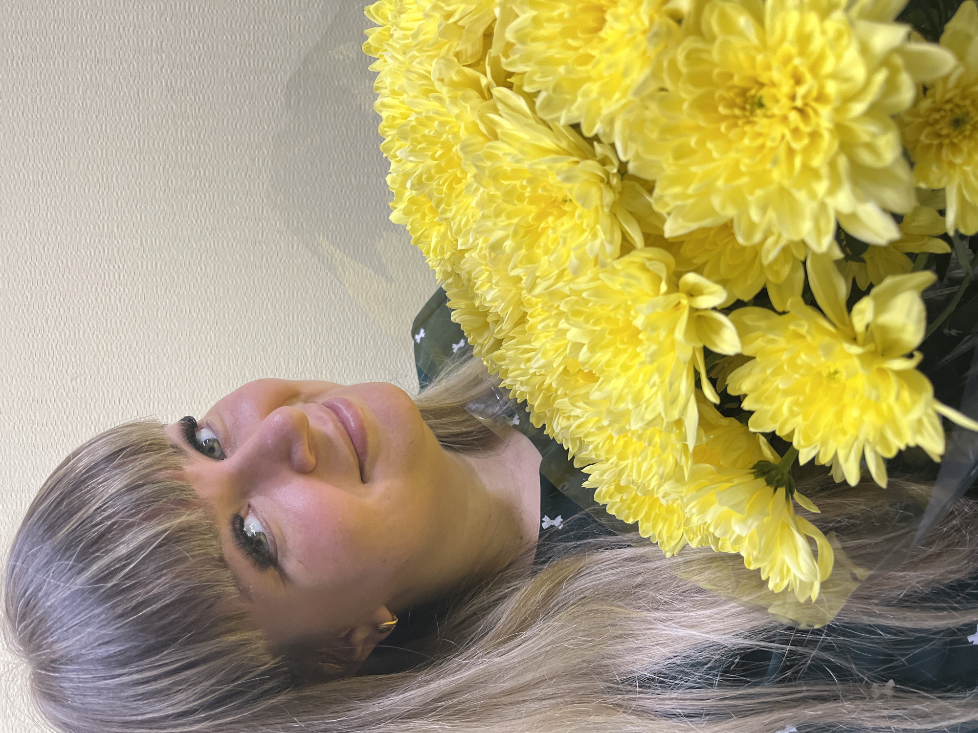 A blonde woman is featured in a portrait-style image, positioned on the left side of the frame, holding a large bouquet of yellow chrysanthemums. The background is a light beige or off-white wall. The woman has fair skin, light blonde hair with bangs, and is wearing earrings. She appears to be smiling subtly. The bouquet of flowers dominates the right side of the image and is densely packed. The image appears to be rotated counter-clockwise. There is no information indicating the location is Izhevsk, Russia.