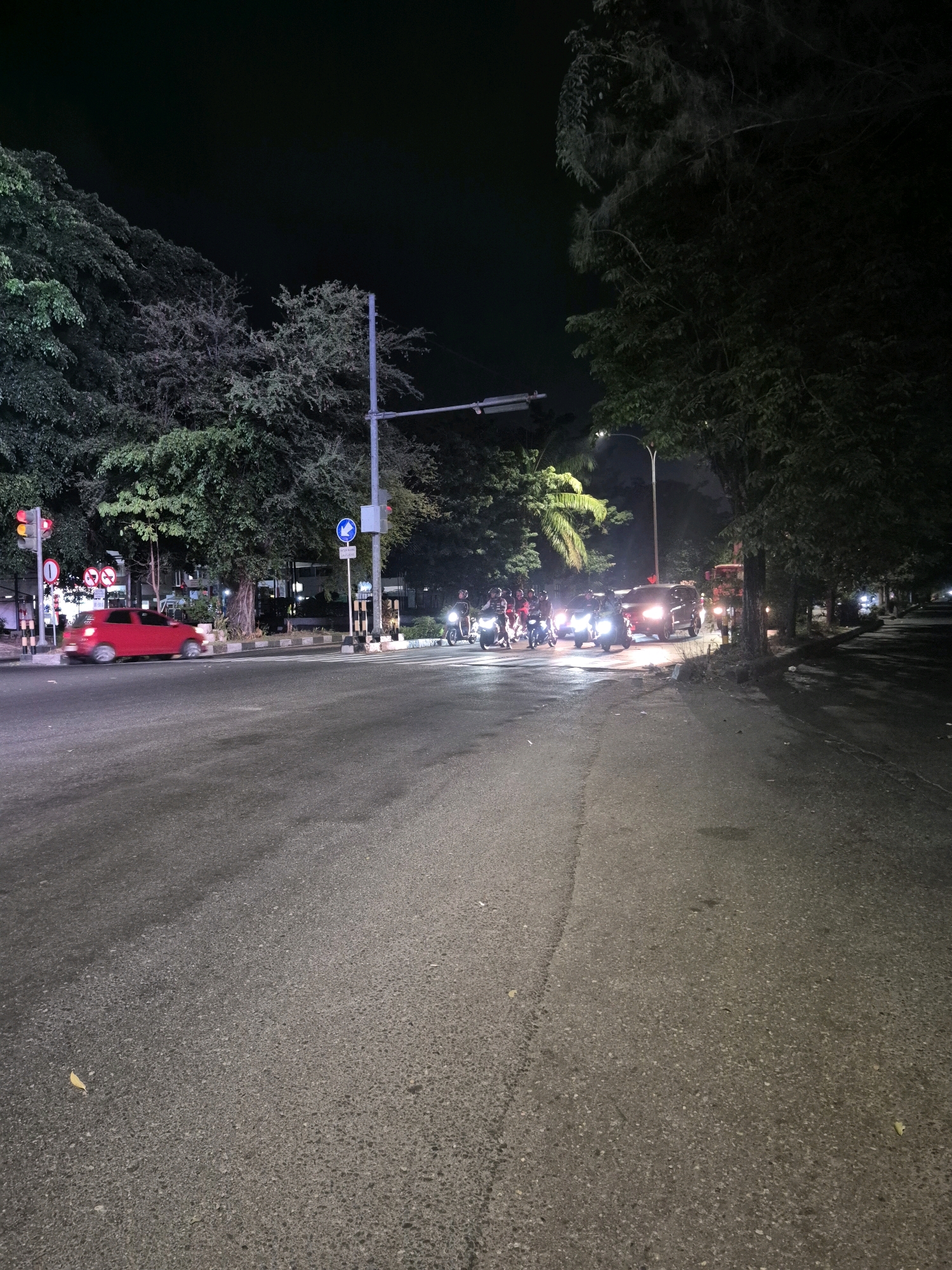 This image depicts a night scene on a city street in Surakarta, Indonesia, likely an intersection. The foreground is dominated by a dark asphalt road with a faint yellow line marking the edge. In the midground, a red car is blurred as it moves from left to right across the intersection. Several motorcycles are stopped or moving slowly on the right side of the road, with their headlights illuminated. A few people are visible on the motorcycles. Traffic lights are present, with one showing red and yellow signals on the left side of the intersection. Signage, including traffic signs and a blue directional sign, is visible along the roadside. Trees line the street, with some foliage illuminated by streetlights. The background is dark, with the sky a deep black, suggesting nighttime. The overall atmosphere is that of a typical urban street scene at night, with activity from vehicles and pedestrians.