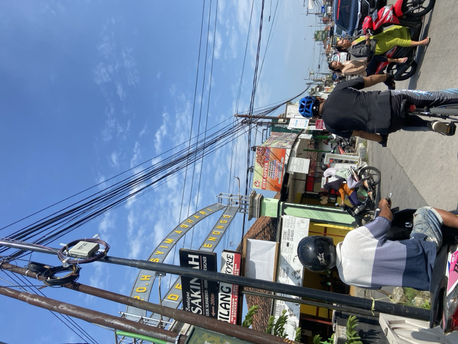 The scene takes place on a street in Kecamatan Tulangan, Indonesia, at the coordinates -7.482 latitude and 112.648 longitude. There are several shops on both sides of the street, with signs indicating the names of the businesses. There are many power lines overhead. Two men are riding bicycles and a group of people are standing around on the sidewalk, including a woman in a blue shirt, a woman in a white shirt, and a woman in a green shirt. In the foreground, a man in a blue shirt and a black helmet is sitting on a pink motorcycle. Two people are riding a motorcycle, and there is a silver car parked on the side of the street. The weather is clear and sunny.