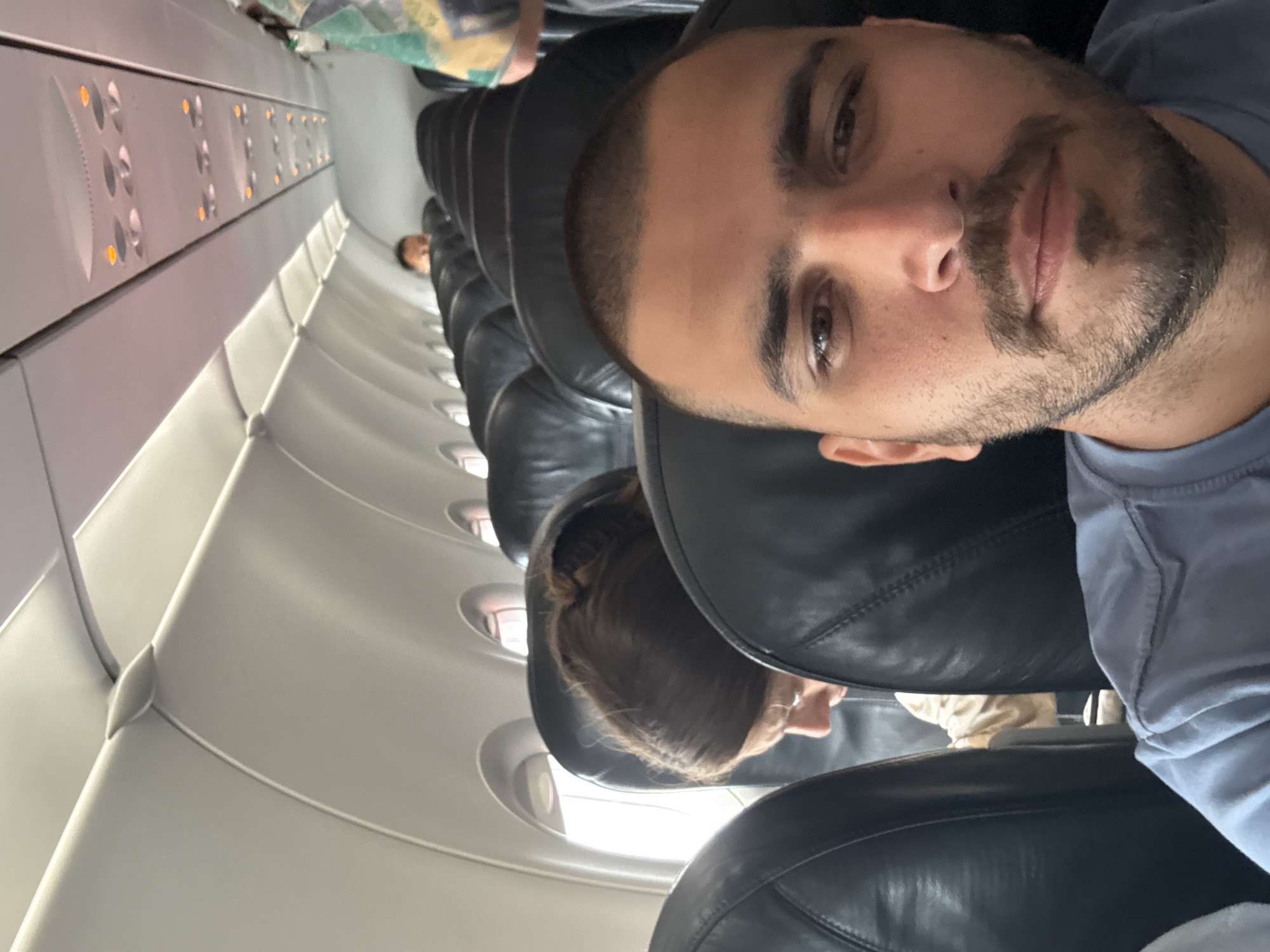 A selfie taken inside an airplane cabin. A man with a beard and short dark hair is in the foreground, facing the camera. Behind him are rows of black leather seats, some occupied by other passengers. The airplane's interior wall is visible on the left. There is no clear evidence in the photo to indicate that this is in the city of Da Nang, Vietnam.