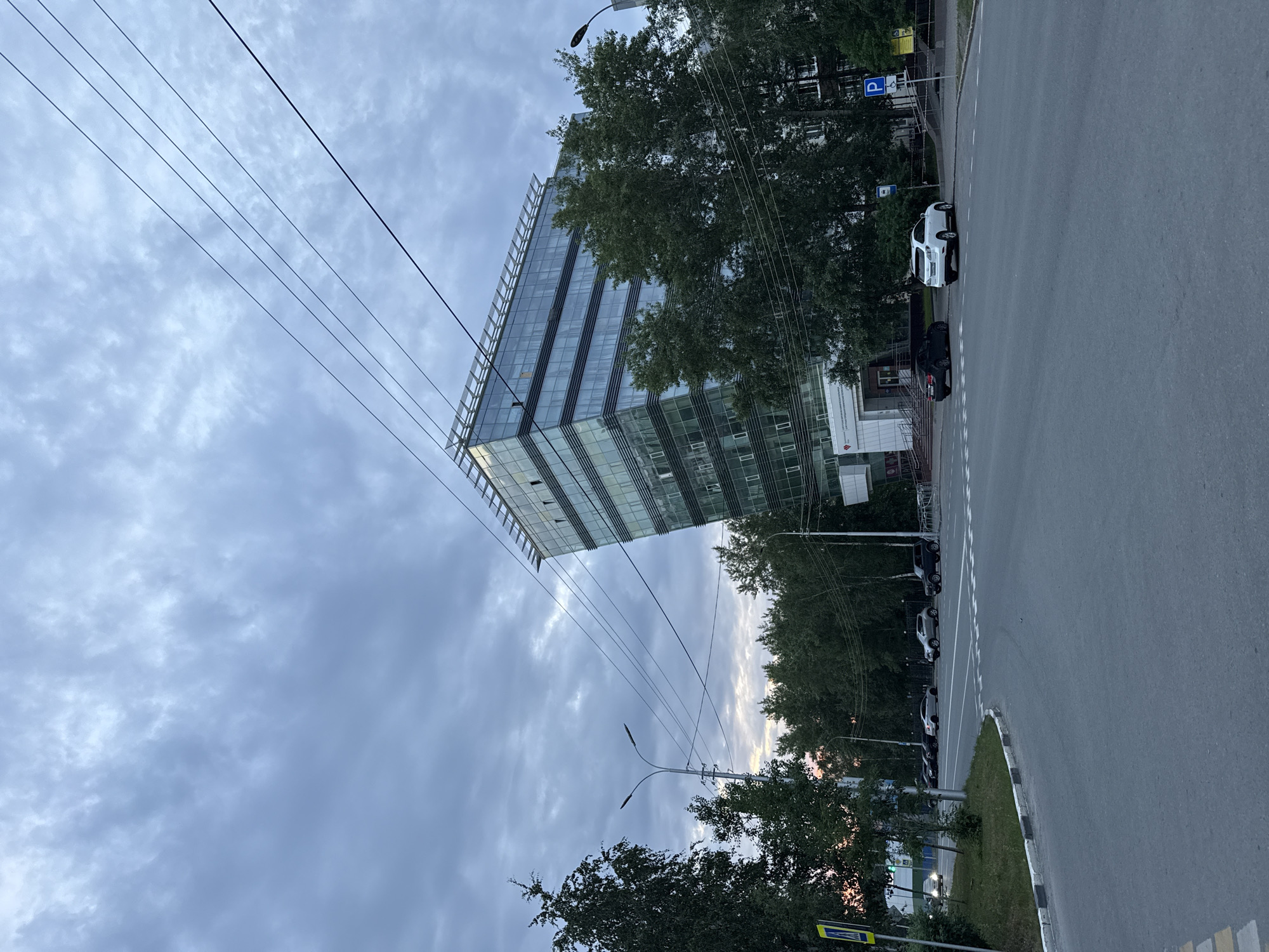 This is a low-angle, outdoor, eye-level shot of a modern glass building in Surgut, Russia. The building, with its sleek, reflective facade, dominates the upper left portion of the frame, its sharp angles cutting into the cloudy sky. Trees with dense green foliage surround the base of the building and line the adjacent street, their branches reaching upwards and partially obscuring the structure. Several power lines crisscross the sky, adding a sense of urban density.
A paved road occupies the right side of the image, with a white and black curb marking its edge. Several cars are parked along the side of the road and the street, including a white SUV and a black sedan. A street lamp with two lights is visible in the lower half of the image, its modern design complementing the contemporary architecture. In the background, through the trees, hints of other buildings and streetlights suggest a developed urban environment. The sky is overcast with dramatic clouds, suggesting either early morning or late evening. There is no clear indication of a specific event, but the scene depicts a typical urban streetscape. Visible text includes a blue parking sign with a "P" symbol and a yellow pedestrian crossing sign.