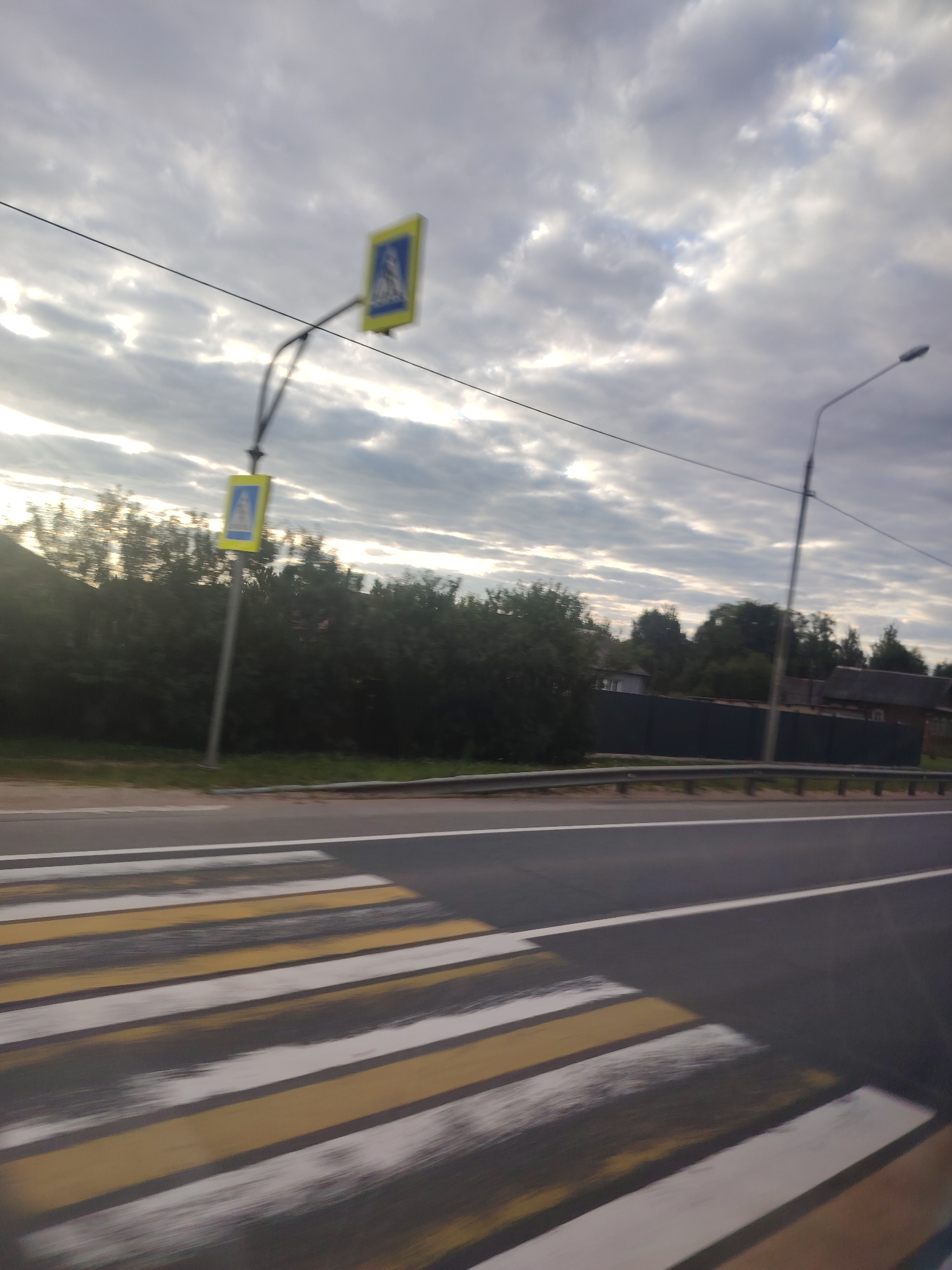 The image shows a pedestrian crossing with yellow and white stripes on a road. A pedestrian crossing sign is visible on a pole to the left of the road, with another smaller sign below it. A street light is also visible on the right side of the road. The background consists of trees and some houses, suggesting a suburban or rural setting. The sky is overcast with clouds. The image was taken in Mokshino, Russia.