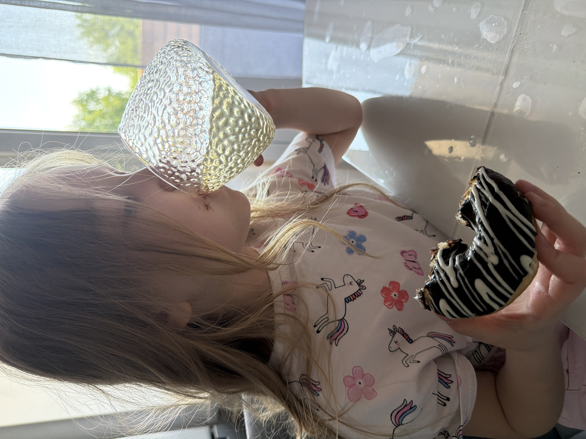 A young child is seated indoors, possibly in a home in Chaikovsky, Russia, based on the location provided. She is drinking from a clear, textured glass with liquid inside. The child is holding a partially eaten, chocolate-glazed donut with white icing in her other hand. She is wearing a white shirt with unicorn and floral patterns. The background appears to be a window with a partially drawn curtain. The light is bright and may indicate daytime. White tiling is visible.