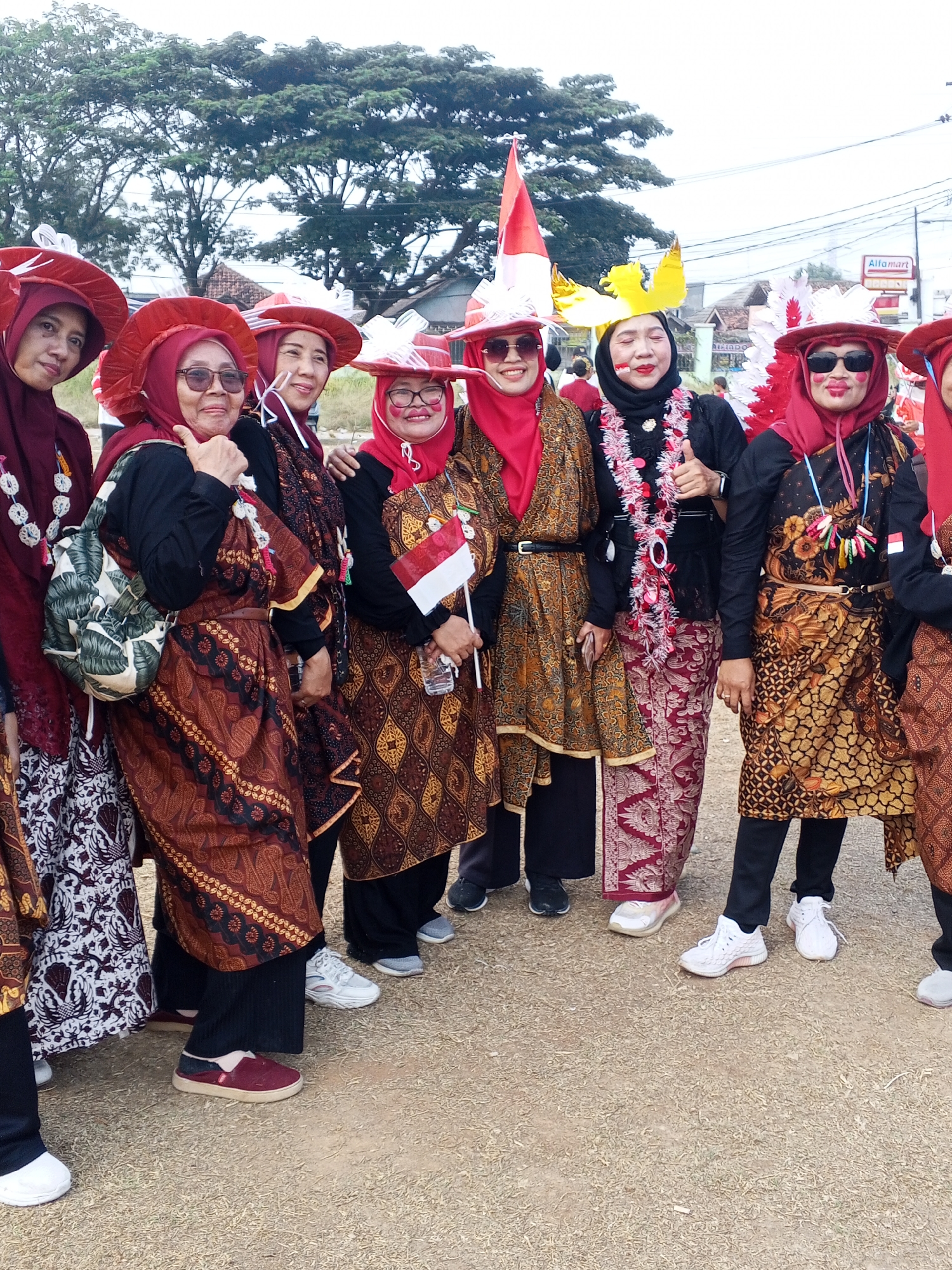In the city of Ciruas, Indonesia, a group of seven women, adorned in festive attire, pose for a photograph. They are positioned outdoors, with a blurred background of trees, buildings, and power lines suggesting a semi-urban or roadside setting. The women are predominantly wearing red headscarves and elaborate, ornate hats, some embellished with feathers and other decorative elements. Their clothing consists of batik-patterned fabrics, including sarongs and blouses, in shades of brown, gold, and red. Several women are wearing necklaces made of beads or festive garlands. One woman holds a small Indonesian flag. Another has a backpack with a leaf-like pattern. The ground beneath them is covered in dry grass or dirt. The overall atmosphere is celebratory, suggesting participation in a local event or festival.