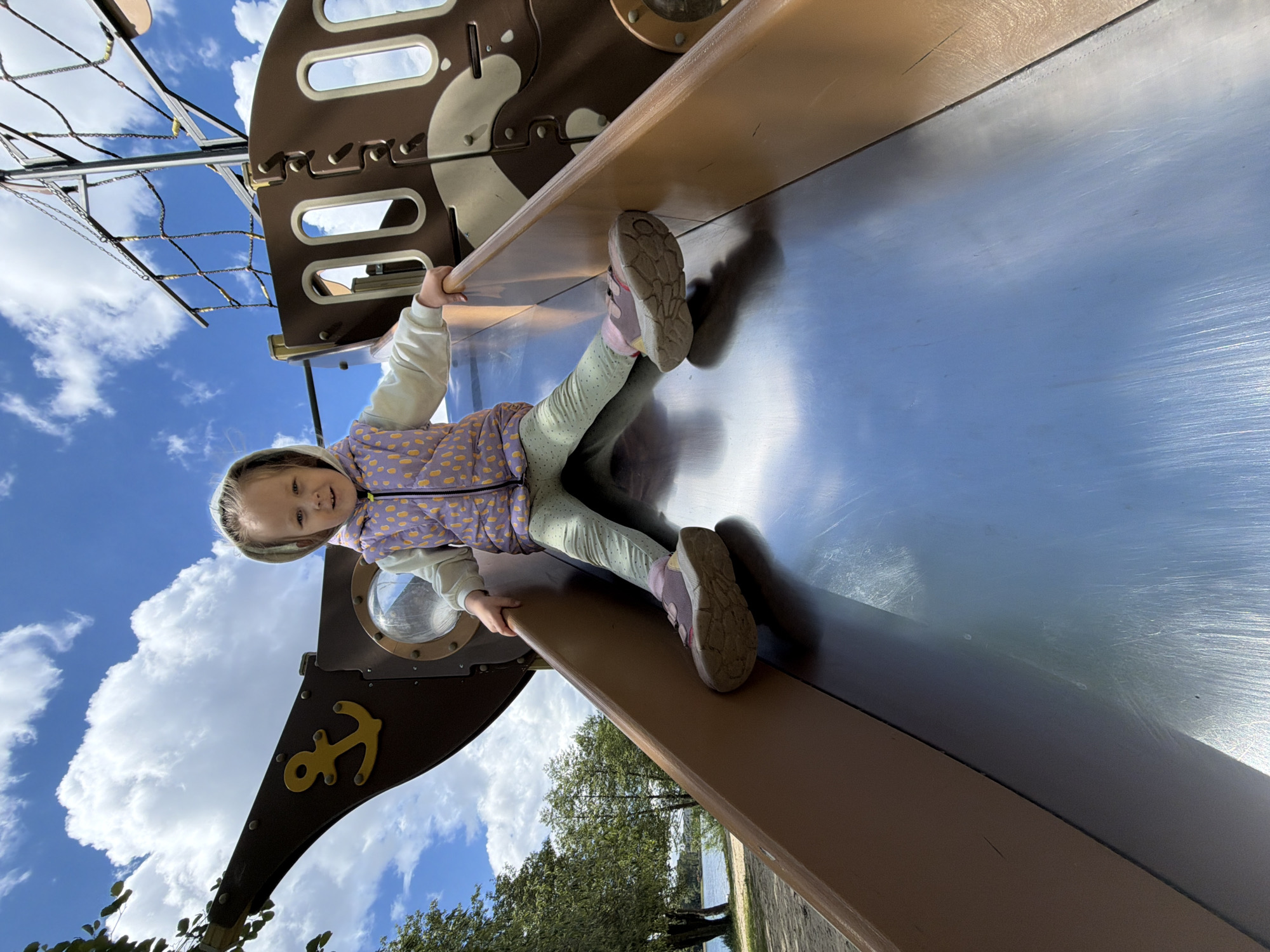 A young girl is on a metal slide that is part of a play structure shaped like a ship. The structure is located in an outdoor area of Malakhovka, Russia. The girl has blonde hair, is wearing a patterned jacket, light green pants, and athletic shoes with pink accents. She is lying on her stomach and holding onto the sides of the slide with her hands. The ship-themed play structure has brown and yellow accents, and the mast includes a rope climbing net. The sky is visible in the background with blue sky and white clouds. Trees and vegetation are visible on the ground. A body of water and tree line is visible in the distance.