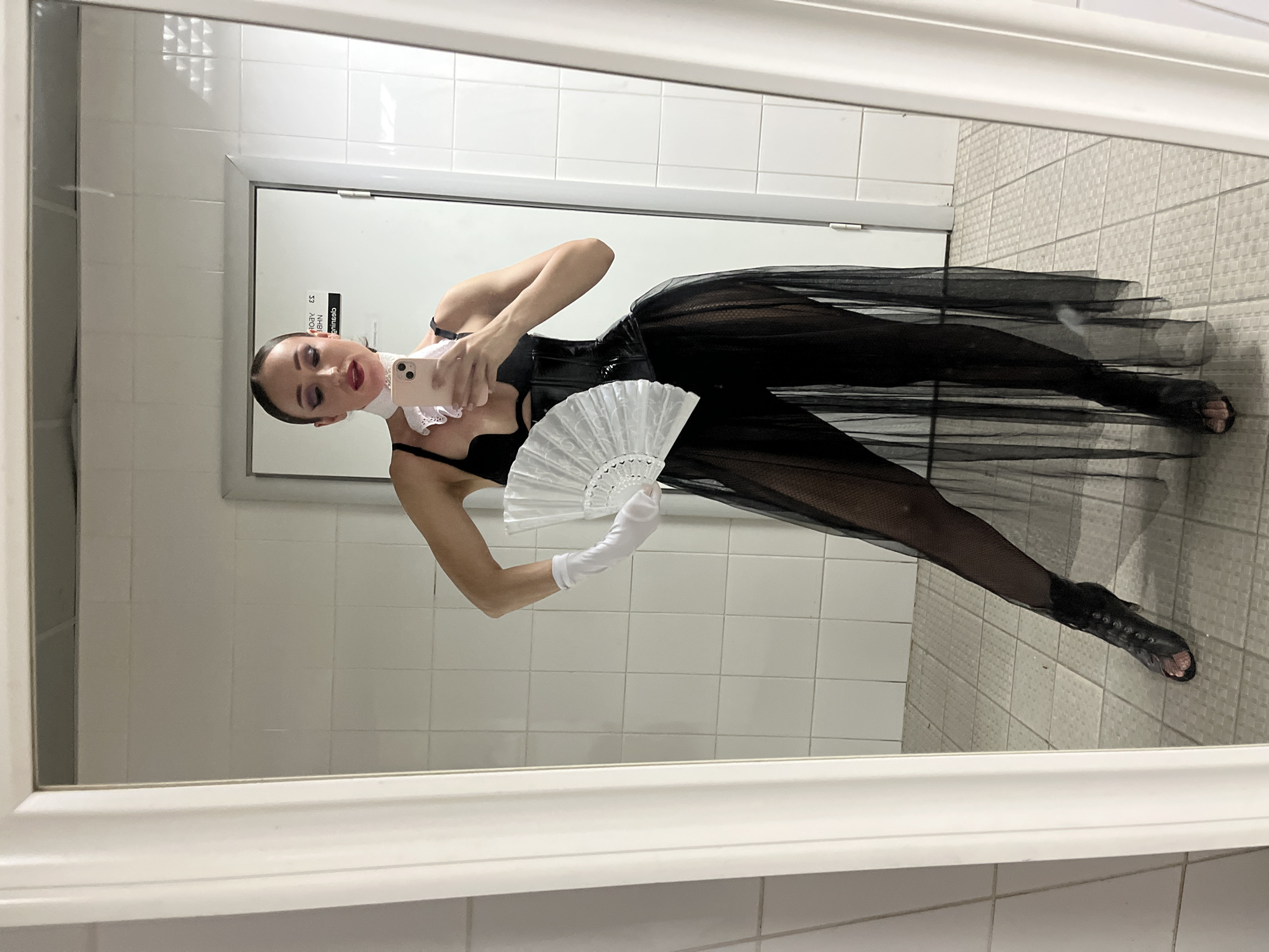 A person is reflected in a mirror, taking a selfie. They are wearing a black corset, a sheer black skirt, white gloves, and black lace-up heeled shoes. A white fan is held in their right hand. The person has dark hair pulled back and is wearing dramatic eye makeup and red lipstick. The setting appears to be a bathroom or dressing room, with white tiled walls and a tiled floor visible in the reflection. A door with a sign is also visible in the background. The lighting is bright, suggesting artificial light. The person's expression is not clearly discernible due to the angle and the reflection.