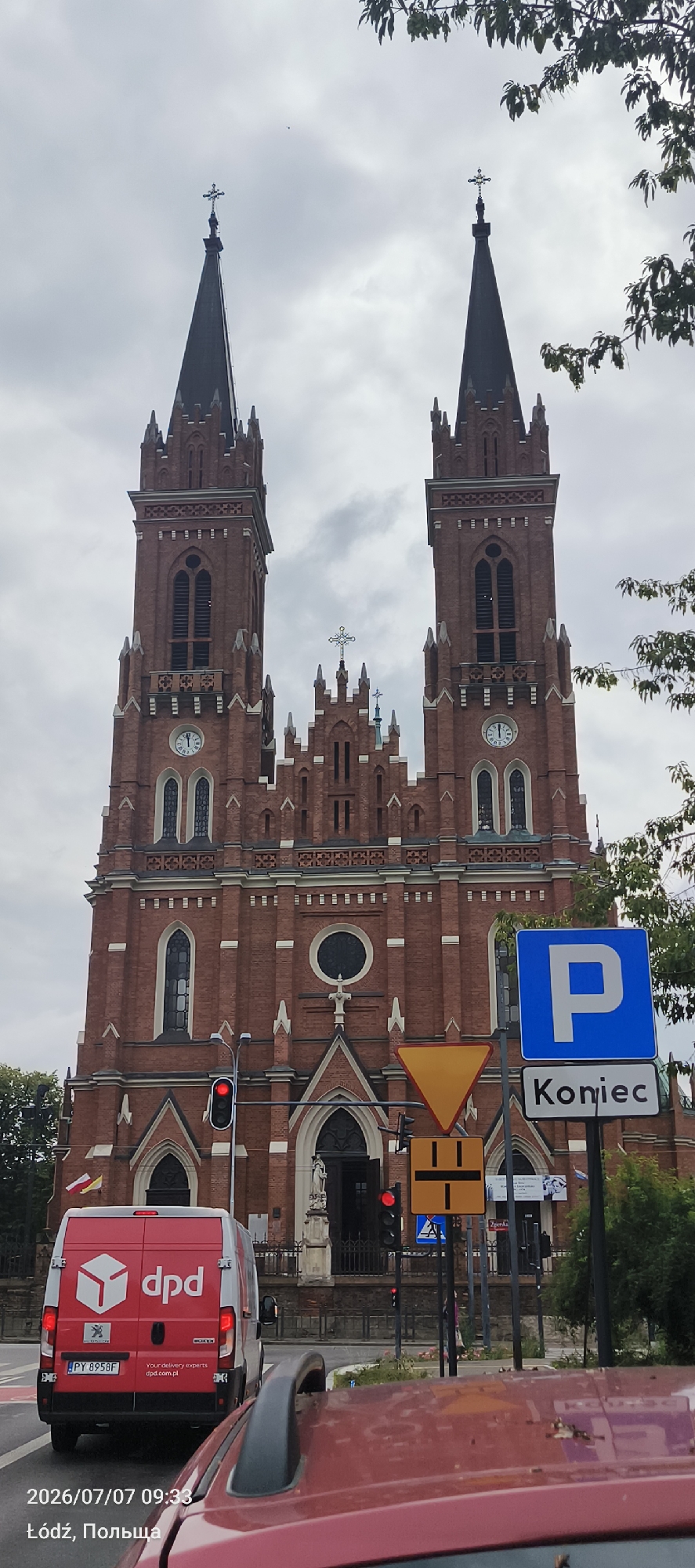 The image shows a red brick church with two tall spires, a classic Gothic Revival architectural style. The church features a large rose window on its facade and a clock on one of its towers. In the foreground, a red delivery van with "dpd" branding is parked, and part of a red car is visible. Traffic lights are present, with one showing a red light. Several street signs are also visible, including a blue parking sign with a white "P" and a white sign below it reading "Koniec" (which means "End" in Polish). The timestamp at the bottom left corner indicates the date and time as "2026/07/07 09:33" and the location as "Łódź, Polska" (Łódź, Poland). The sky is overcast with clouds.