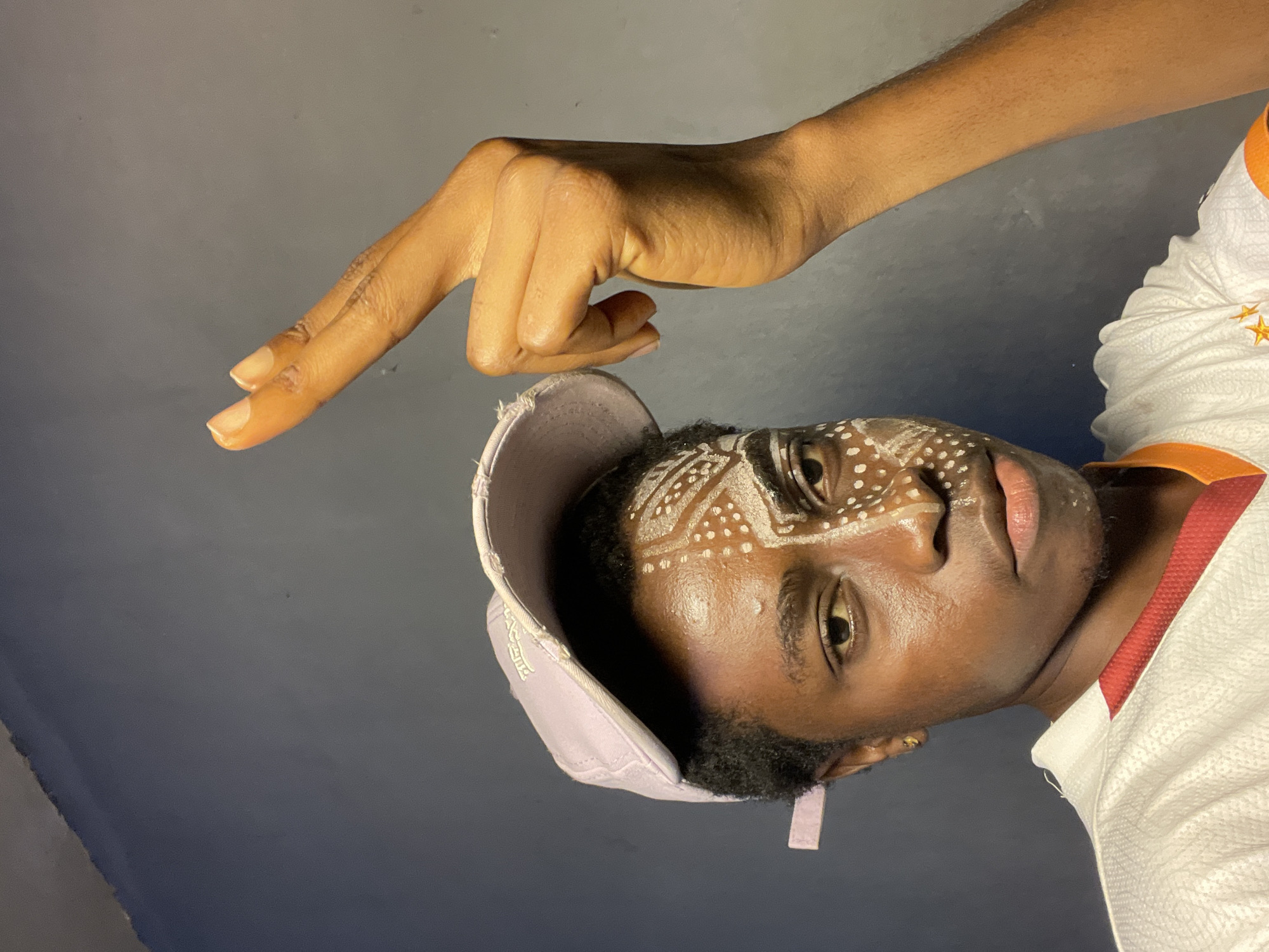 A close-up studio shot shows a dark-skinned man with tribal-style white face paint, wearing a light purple cap, and a white shirt with orange trim. The man's face is angled towards the upper right. His left hand is raised above his head, with the index and middle fingers extended. The background is a dark gray. The location is not evident in the image.