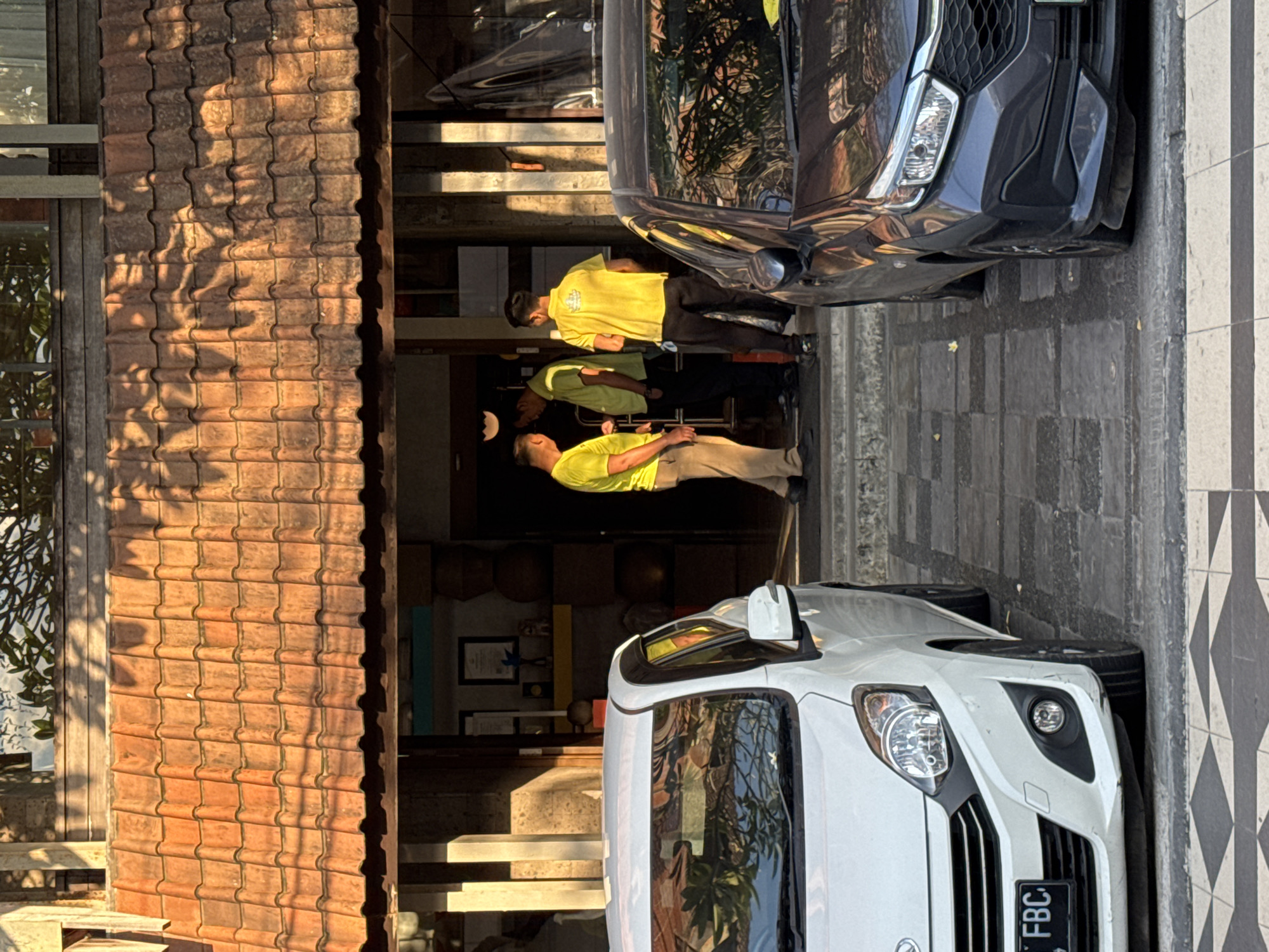 The image captures an outdoor scene in Kuta, Indonesia, featuring the entrance of a building with three men and two parked cars under bright, direct sunlight. The scene is likely a street or driveway in front of a commercial establishment, such as a shop or hotel.
Three men, appearing to be staff members, are positioned at the building's entrance. They are all wearing bright yellow short-sleeved shirts, indicative of a uniform. Two men wear dark trousers, while the third wears light-colored trousers. One man is leaning against a wooden pillar on the left side of the entrance, with another standing slightly behind him. The third man is positioned further inside the doorway. Their relaxed postures suggest they are waiting, observing, or engaged in a casual discussion.
Two cars are parked parallel to the building on a paved surface. The car in the foreground on the right is white, with its front fascia, headlights, and part of its grille clearly visible. Its license plate displays the text "FBC". Behind it, a dark gray or black sedan is parked, showing its front bumper and headlights. The cars' glossy surfaces reflect the surrounding environment, including foliage and parts of the building.
The building features a distinctive reddish-brown tiled roof with a ridged pattern, on which sharp, intricate shadows of unseen foliage are cast, highlighting the strong sunlight. Wooden architectural elements, such as window frames and pillars, are also visible. The ground consists of gray paving stones, transitioning to a section of black and white checkered tiles on the far right. The strong, directional shadows on the roof and pavement indicate the time of day is either morning or late afternoon, under clear, sunny weather.