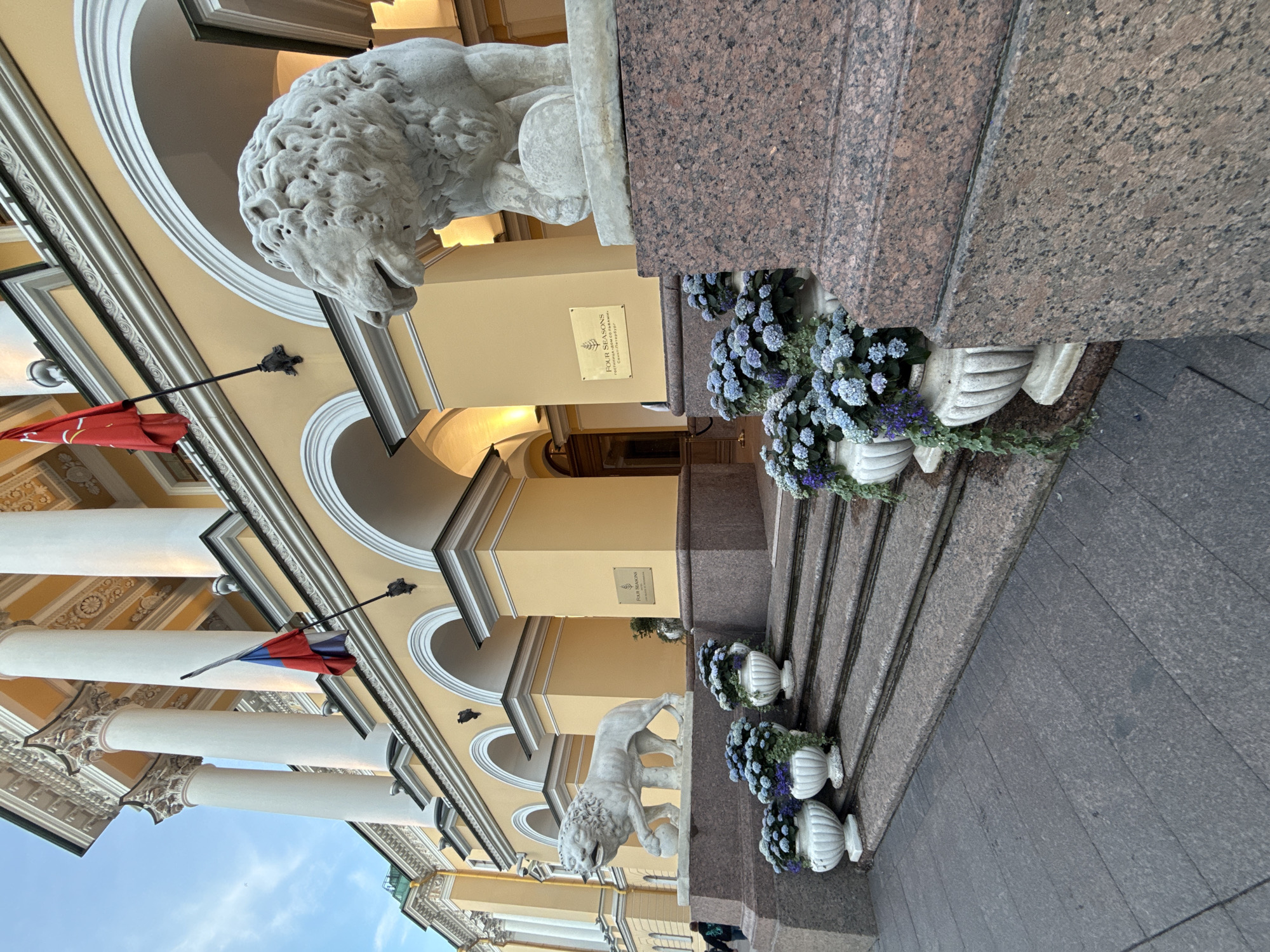 The image shows the entrance to the Four Seasons Hotel Lion Palace in Saint Petersburg, Russia. Two large white stone lions flank the entrance, with the one on the right being closer to the camera. The building is painted a pale yellow and features grand columns and arched doorways. Russian flags are visible on poles near the top of the entrance. A set of granite stairs leads up to the entrance, decorated with white planters filled with blue and purple flowers. The ground in front of the stairs is paved with dark grey stone. In the background, a person can be seen walking away from the camera on the left side of the image. The sky is visible at the top, with a hint of twilight.