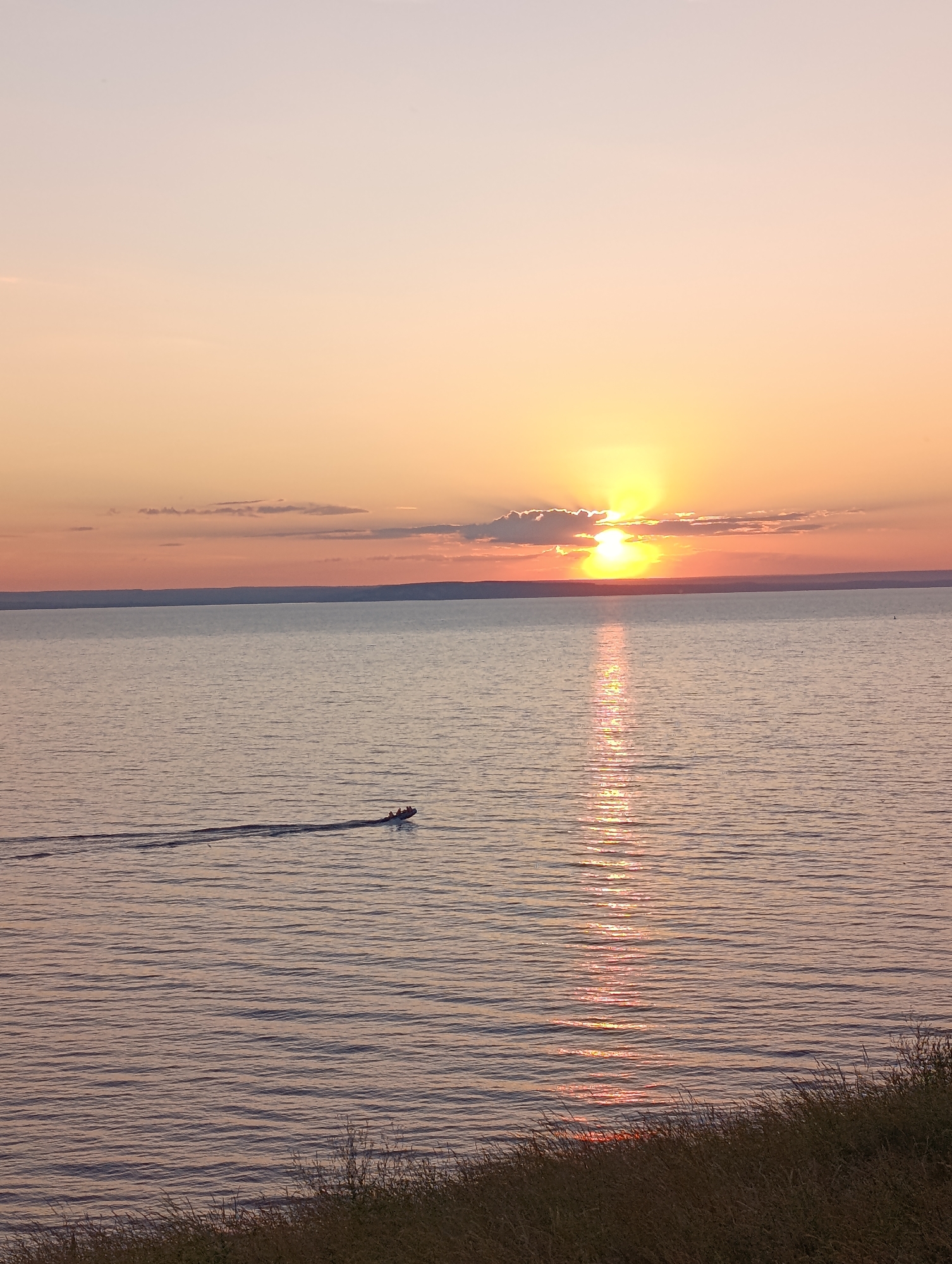 An eye-level, vertical shot captures a serene body of water at sunset. The sun, partially obscured by a cloud, casts a warm, golden glow across the sky and reflects vibrantly on the water's surface. A small motorboat with several occupants is seen in the distance, leaving a wake as it moves across the tranquil sea. The foreground features a grassy embankment, adding a natural touch to the scene. The location is Unknown, Unknown.