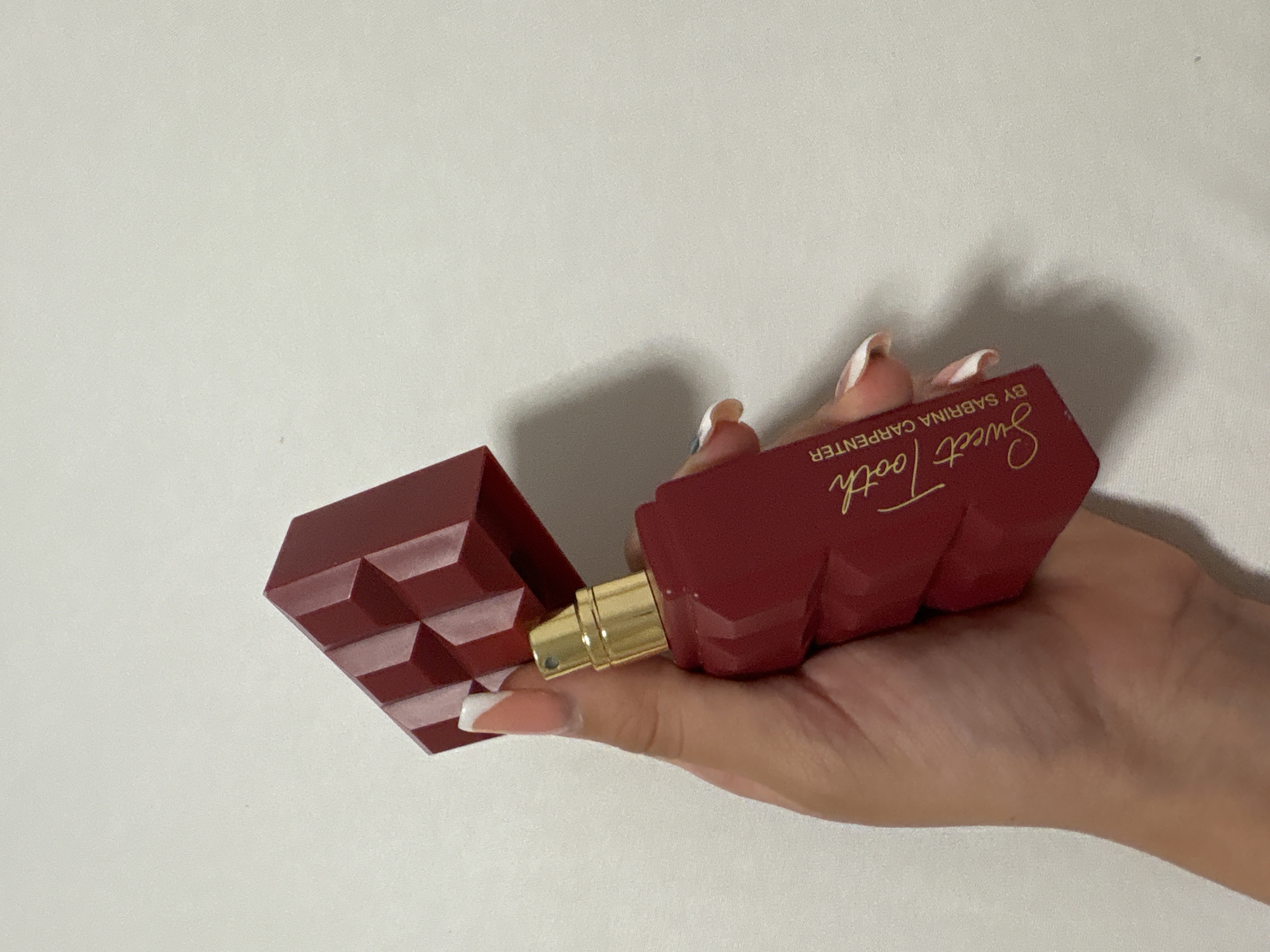 A person's hand is holding a red perfume bottle with a gold spray nozzle. The bottle has a chocolate bar-like design and gold text that reads "Sweet Tooth by Sabrina Carpenter." The background is a plain white wall.