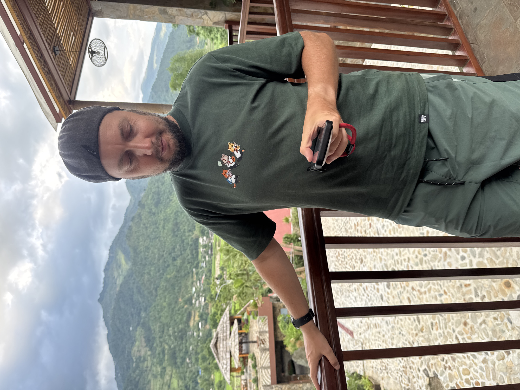 A man with a beard, wearing a gray cap, green t-shirt with cartoon figures, and green pants, stands on a balcony overlooking a valley. He holds a black phone with a red lanyard in his right hand and rests his left hand on a wooden railing. The balcony has a stone floor. In the background are mountains, a cloudy sky, and a village with thatched-roof structures. The location is likely in or near Tú Lệ, Vietnam, based on the typical scenery.