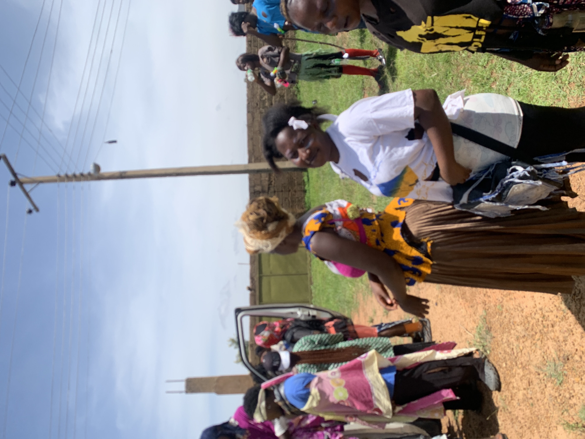 The image shows a group of people outdoors, possibly in Gwafan, Nigeria. The setting appears to be a rural or semi-rural area with a dirt ground, grass, a brick wall in the background, and a utility pole with overhead wires. Several individuals are visible, dressed in various colorful clothing and head coverings. Some are standing, while others appear to be sitting or moving. One person in the foreground is looking towards the camera. The overall impression is of a gathering or event taking place in an open-air location.