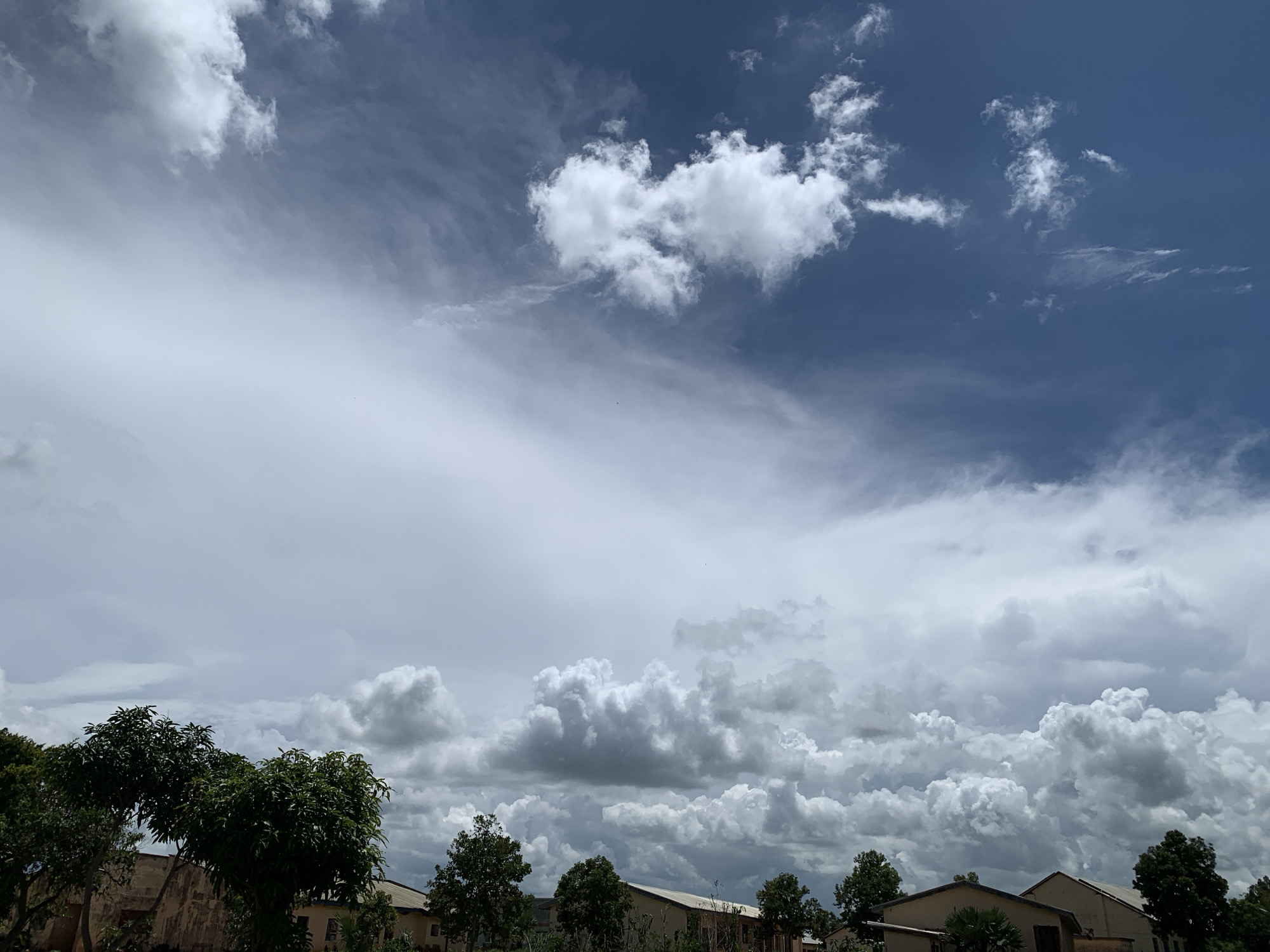 The image depicts a cloudy sky over a residential area in Kuru Karama, Nigeria. The sky is a mix of dark and light clouds, with a deep blue sky visible in the upper right corner. Below the sky, several buildings with tan exteriors and dark roofs are visible, interspersed with lush green trees. The foreground features more trees and foliage. The scene appears to be captured during daylight hours.