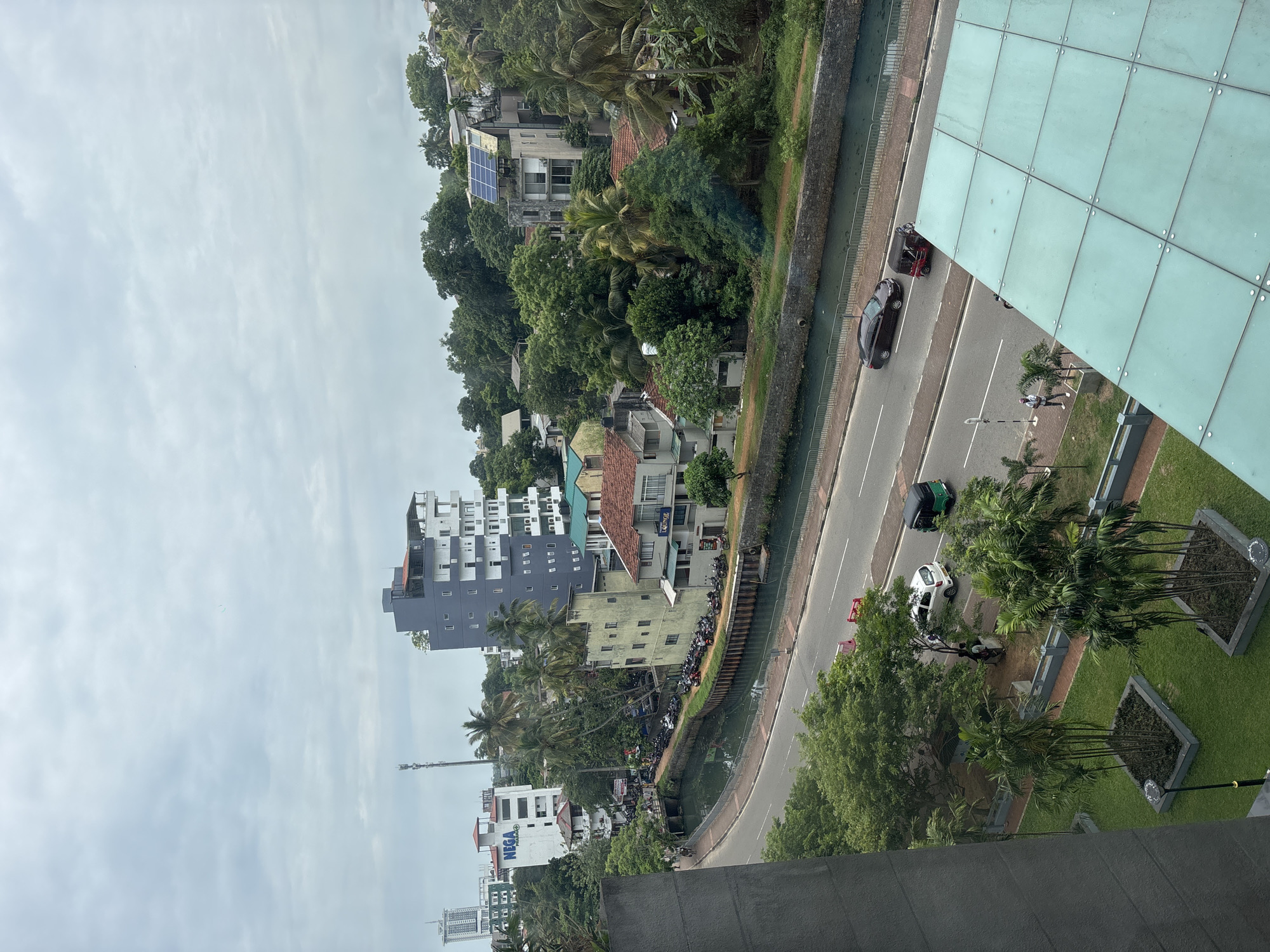 A high-angle, daytime view shows a section of Battaramulla, Sri Lanka. The scene includes buildings, a road with vehicles, a canal, and greenery. Buildings range in height and architectural style, from multi-story apartments to smaller residential homes. The road is adjacent to the canal, with vehicles including cars and a three-wheeled auto-rickshaw (tuk-tuk). The canal runs parallel to the road, with vegetation along its banks. Trees and green spaces are dispersed throughout the view. People are visible along the edge of the road, walking. The sky is overcast. A portion of a modern glass building is visible on the right side of the image, framing the scene.