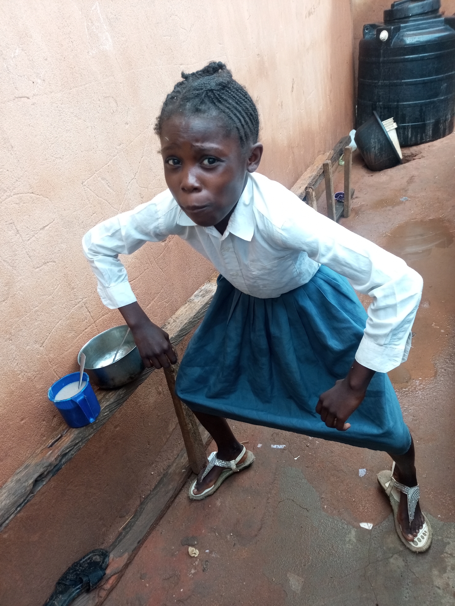 A young Black girl, appearing to be of school age, stands with her knees bent and arms outstretched in a crouched position. She is wearing a white button-up shirt and a blue skirt. Her hair is styled in braids. In front of her, to the left, is a blue plastic mug filled with a white liquid, and a metal bowl also containing a white substance, possibly food. To the right and in the background, a large black water tank is visible. The ground is a reddish-brown, wet surface, indicating it may have rained. The wall behind the girl is a textured, light-colored surface. The scene appears to be an outdoor area, possibly a yard or alleyway, with a rustic wooden structure to the right. The overall impression is one of a candid shot, capturing the girl in a dynamic pose in a simple, utilitarian environment. The location is identified as Tempure, Nigeria.