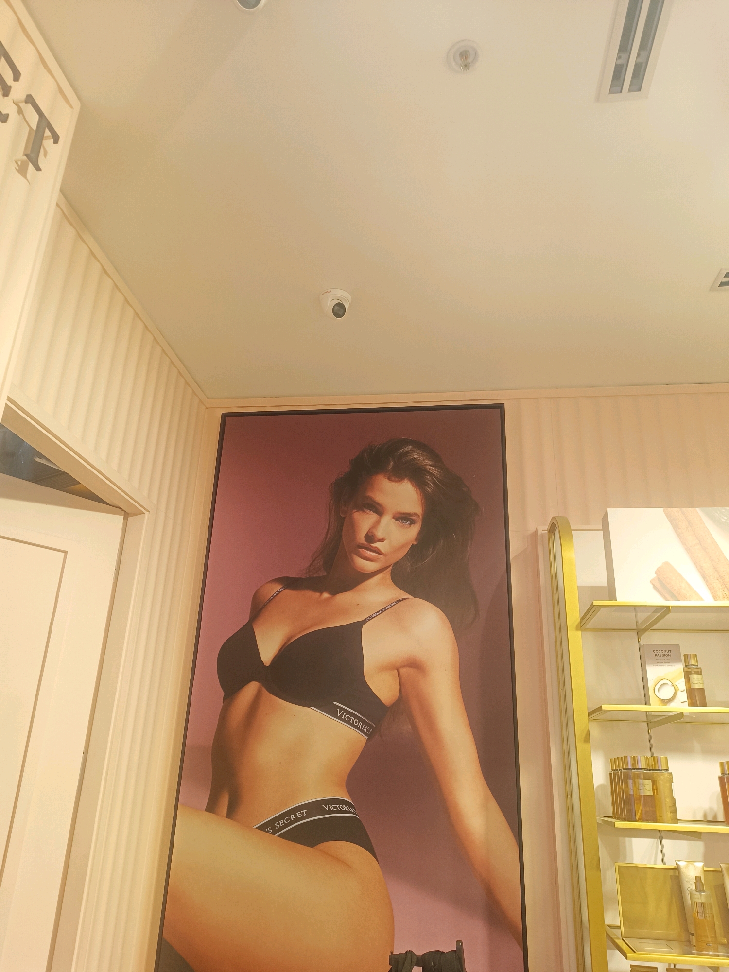 The image depicts a retail display within a Victoria's Secret store. Dominating the center is a large poster of a female model wearing black lingerie, the "Victoria's Secret" logo visible on the waistband of her underwear and bra. To the left, a cream-colored doorframe and wall paneling suggest a fitting room or entrance. To the right, a gold-colored shelving unit displays various perfume bottles and cosmetic products. The ceiling features recessed lighting and a security camera. The overall lighting is warm and natural, possibly indicating the time of day. There is no indication of the specific location within Hyderabad, India, as the image only captures the interior of the store.