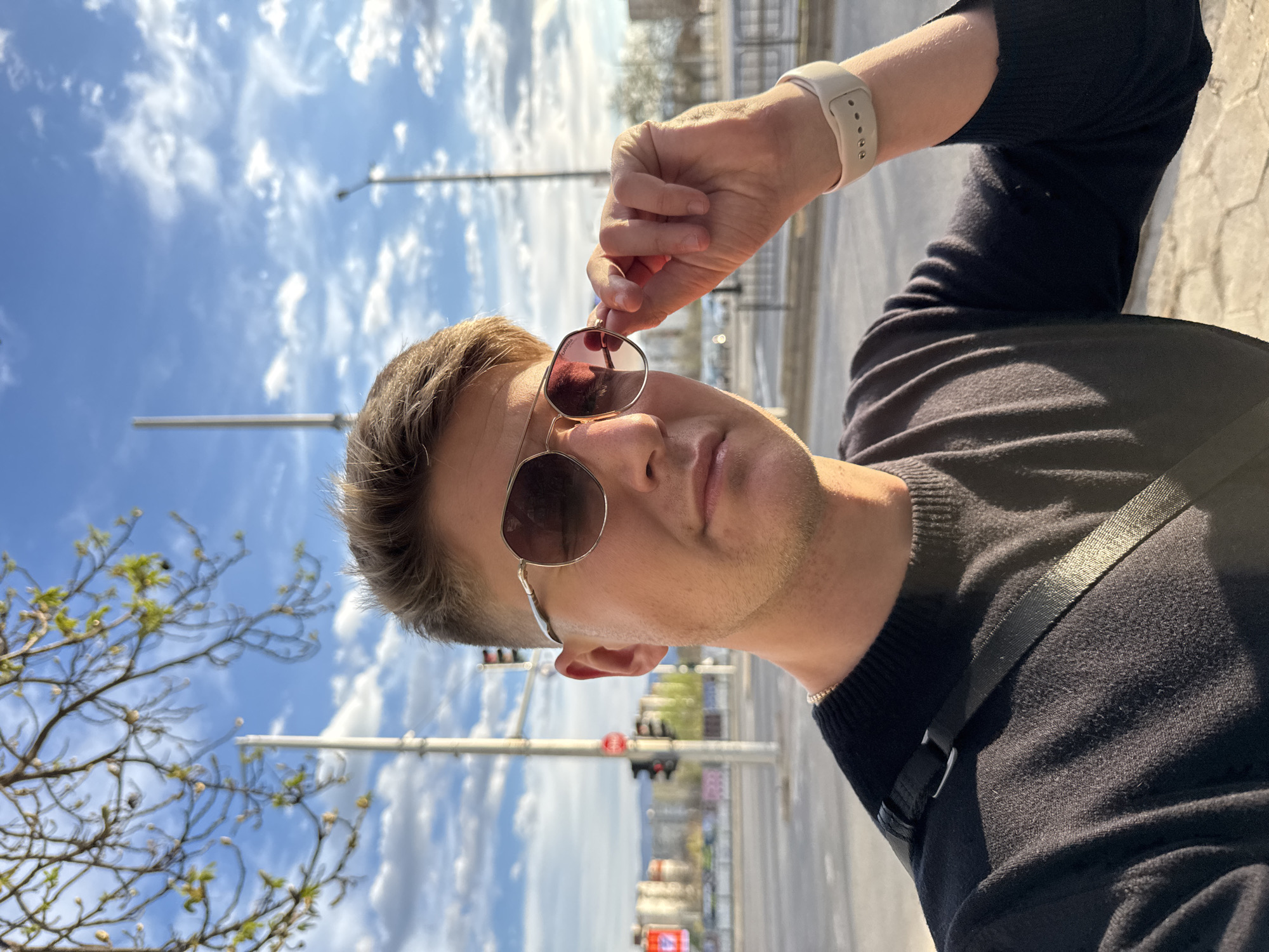 A man is holding his sunglasses up to his face. He has short, light brown hair and is wearing a black sweater. He has a watch on his wrist and a strap of some kind across his chest. Behind him is a street scene with traffic lights and other signage. There are trees on the left side of the frame. The sky is blue with white clouds.
The location is Plovdiv, Bulgaria.