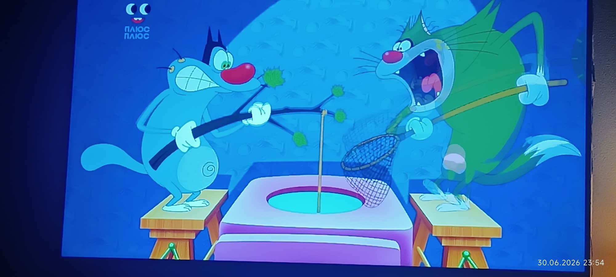This image is a screenshot from a cartoon featuring two anthropomorphic cats engaged in an activity. On the left is a light blue cat holding a stick with green fuzzy objects attached. On the right is a green cat with its mouth wide open, holding a net and a stick. They appear to be interacting with a purple object that has a circular opening. The background is a solid blue with a subtle textured pattern. In the upper left corner, there is a logo with two circles and text that reads "ПЛЮС ПЛЮС". In the lower right corner, a timestamp "30.06.2026 23:54" is visible. There is no indication of the city of Odesa, Ukraine, or any specific urban environment.