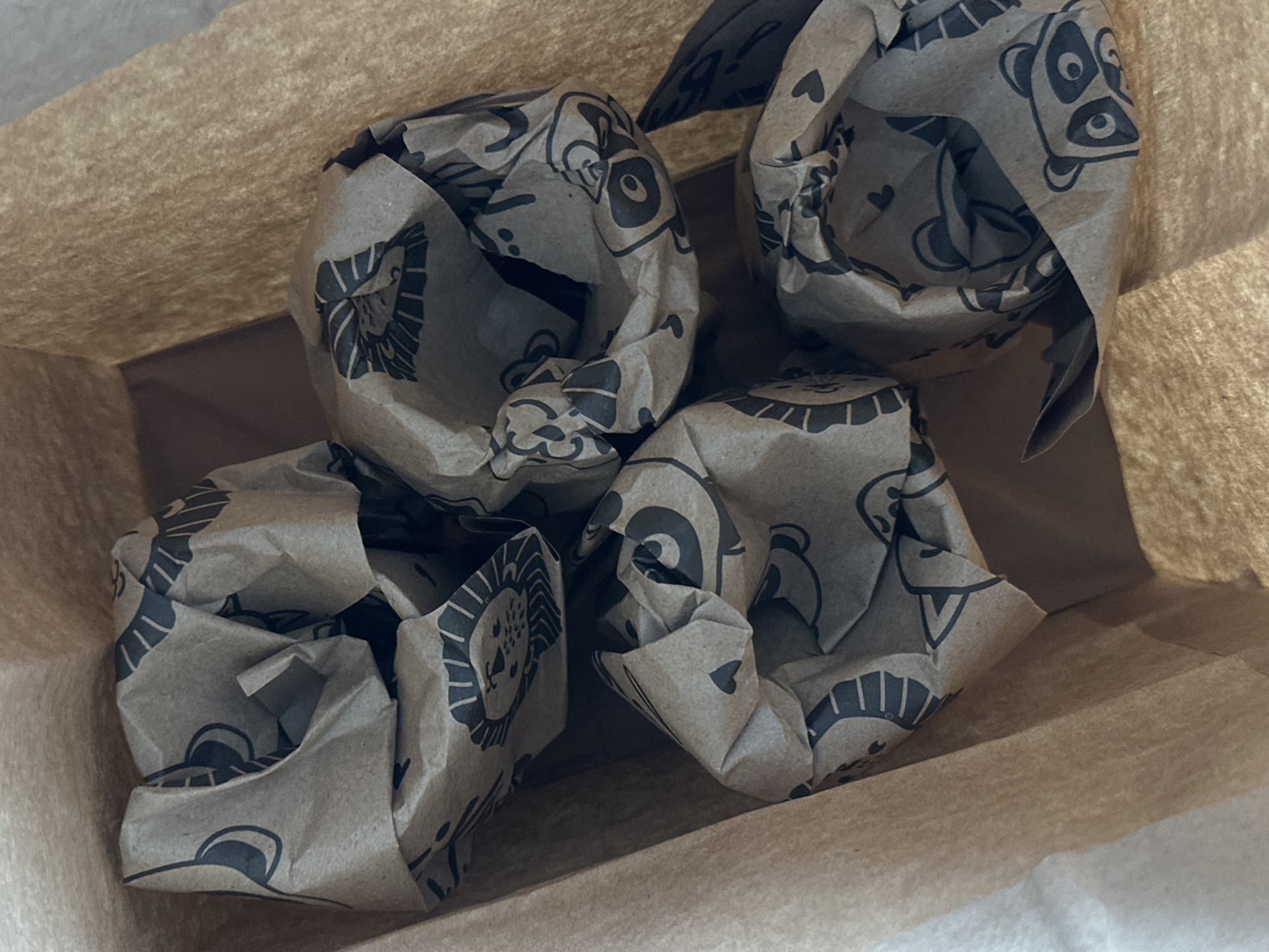 The image presents a close-up, overhead view looking into a light brown paper bag. Inside the bag are multiple crumpled pieces of darker brown paper, which appear to be wrapping paper or tissue paper. These pieces of paper are adorned with a repeating pattern of black, cartoon-style line illustrations.
The visible illustrations include distinct animal faces such as pandas, characterized by their large eyes and dark patches, and lions with their manes. Other less defined animal caricatures and small heart shapes are also scattered throughout the pattern. The paper is heavily creased and folded, creating depth and texture within the bag. The main paper bag itself shows some visible folds and a slightly fibrous texture, characteristic of kraft paper. The background within the bag's deeper sections appears dark brown or grey, indicating shadow or the bag's interior base.
There is no activity or event explicitly taking place; the image captures a static collection of items within the bag. The lighting is soft and diffuse, offering no specific clues about the time of day or outdoor weather. No people are visible, nor are there any discernible emotions conveyed. Aside from the animal and heart patterns, no readable text is present on the paper. While the location context is provided as Kuda, Russia, the image itself does not contain any visual cues specific to this geographical location.