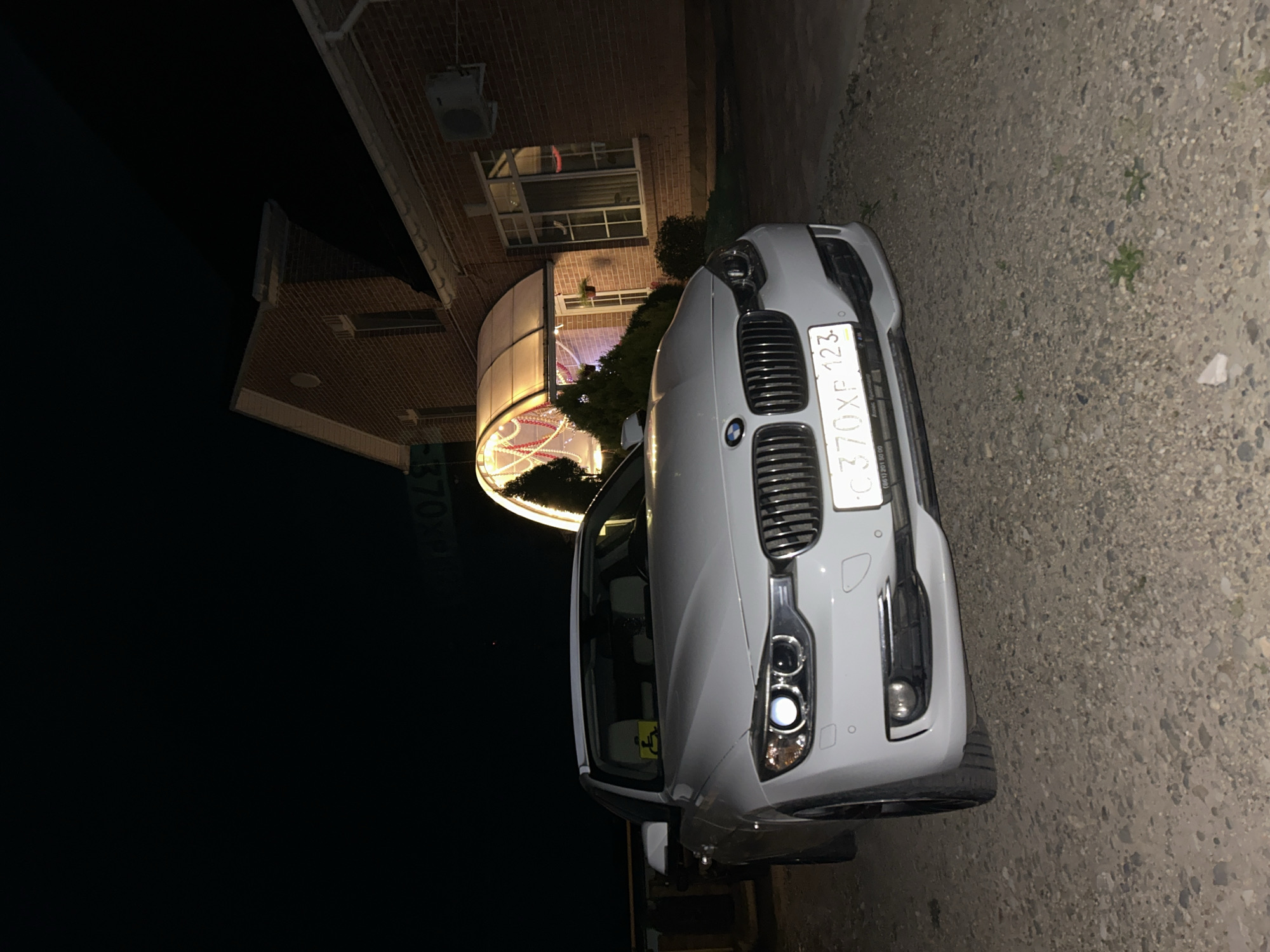 A white BMW SUV is parked on a gravel driveway at night, in front of a brick building with a large, illuminated curved window. The building has a covered entrance with lights and decorations, possibly for a holiday. The car's headlights are on, illuminating the road and the front of the vehicle. The license plate is visible, reading "C370XP 123". The scene appears to be in a suburban or rural area, given the gravel and the dark sky. The overall mood is calm and still, suggesting a quiet evening. The image is a still photograph taken from a low angle, showing the front of the car and the building.