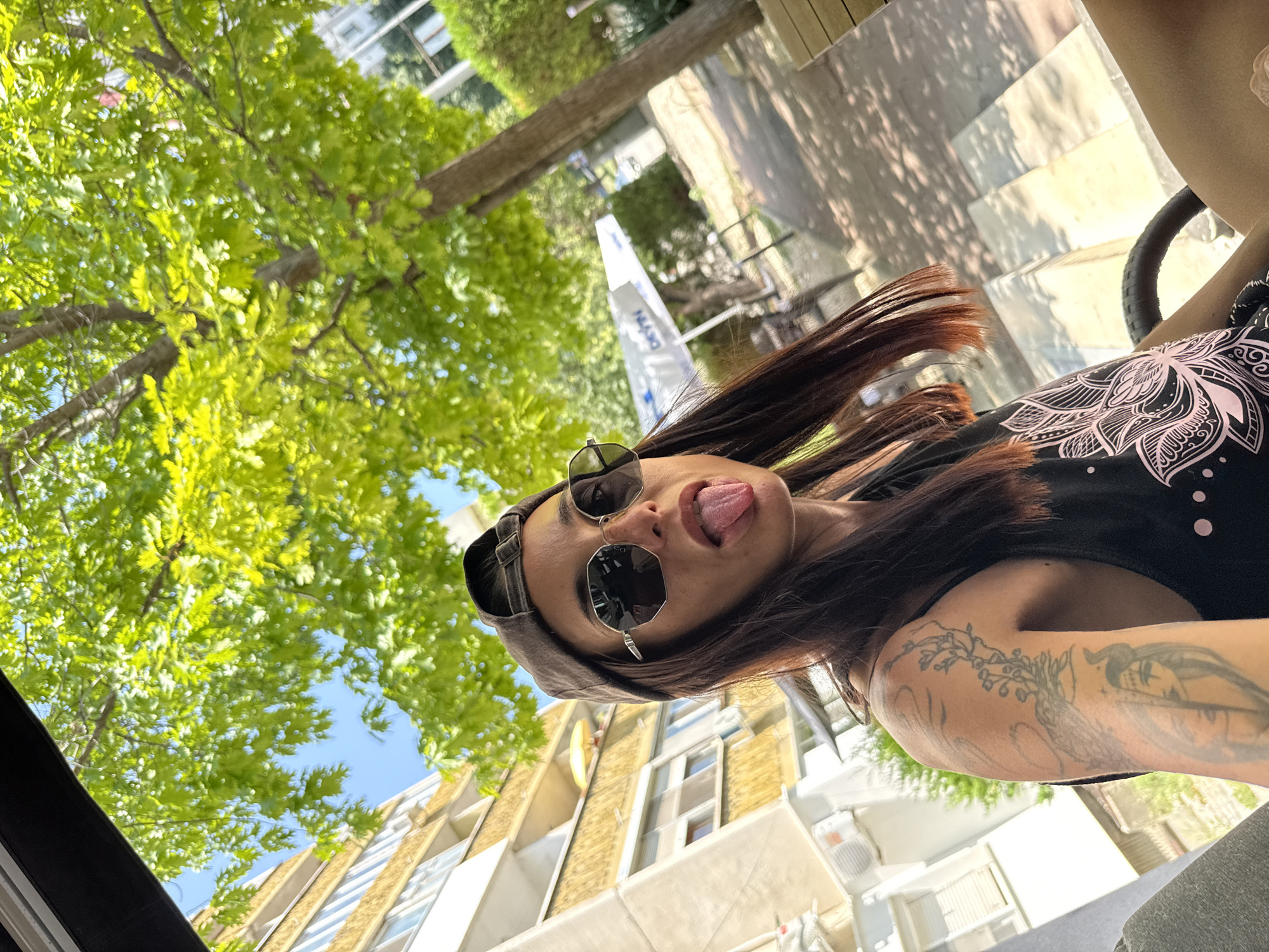 A young woman with long brown hair, wearing a baseball cap and sunglasses, sticks her tongue out at the camera in a selfie taken outdoors in front of a building. She has tattoos on her left arm and is wearing a black tank top with a pink floral design. In the background, a multi-story building, trees, and outdoor seating are visible. The location appears to be a cafe or restaurant in the city of Pleven, Bulgaria.