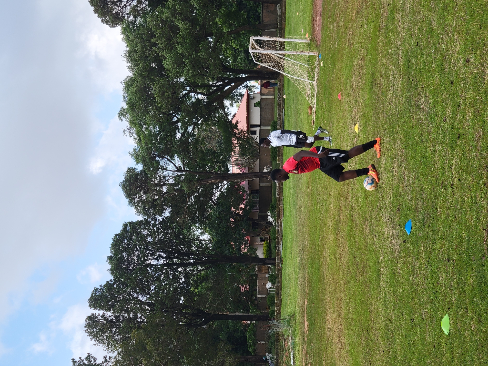The image depicts a football training session on a grassy field. Two young men, dressed in athletic wear, are engaged in practice. One player, wearing a red shirt and black shorts, is in the foreground with a soccer ball at his feet. The other player, in a white shirt and black shorts, is positioned further back. A soccer goal with a net is visible in the background, along with several colorful training cones scattered across the field.
The setting appears to be an outdoor sports field, possibly a school or club facility, with lush green grass. Trees with dense foliage surround the field, and buildings with red-tiled roofs are visible behind the trees, suggesting a semi-urban or suburban environment. The sky is partially cloudy, indicating it is daytime, likely late morning or afternoon, with fair weather.
The overall atmosphere suggests a focused training session, with the athletes concentrating on their drills. There is no visible text in the image. The scene is set in Jos, Nigeria.
