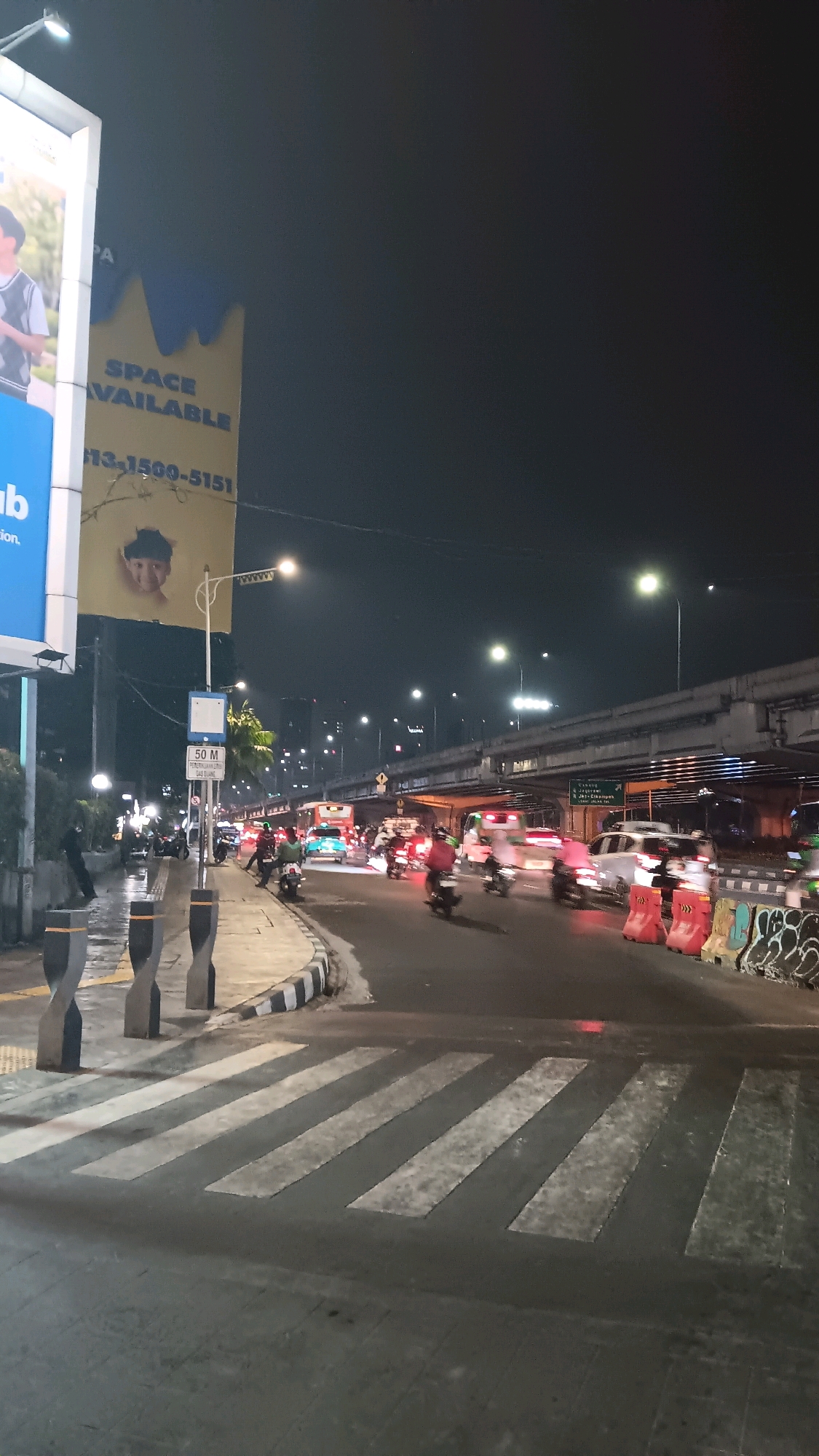 The image captures a nighttime street scene in South Jakarta, Indonesia, likely at an intersection. The foreground features a wet pedestrian crossing with prominent white stripes. To the left, a sidewalk is lined with decorative, twisted bollards and a bus stop shelter with a sign indicating "50 M". Beyond the sidewalk, a large billboard is visible, part of which displays text like "SPACE AVAILABLE" and a phone number. The background is dominated by a multi-level overpass with vehicles, including cars and motorcycles, moving along the road below and on the elevated structure. The lighting suggests urban nocturnal activity, with streetlights, vehicle headlights, and illuminated buildings contributing to the scene's ambiance. The overall impression is one of a busy urban environment during nighttime hours.