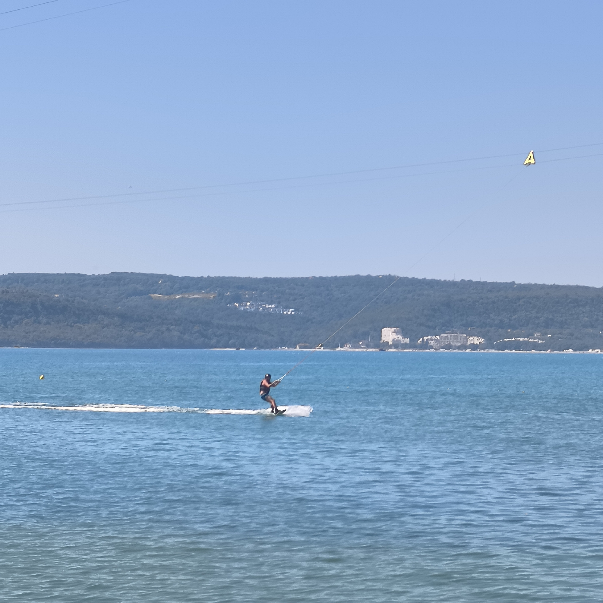 The image shows a person kiteboarding on the Black Sea near the city of Varna, Bulgaria. The kiteboarder is in the center of the frame, wearing a dark top and blue shorts, holding onto a kiteboarding bar. They are riding on a white board that is leaving a white wake behind them. The water is a vibrant blue, with small waves. In the background, a lush green, tree-covered hill rises from the coastline. On the hillside, several buildings, including what appears to be a large white hotel or resort, are visible. The sky is a clear, bright blue, with thin power lines stretching across the upper part of the frame. One of the power lines has a yellow triangular marker with the letter "A" on it. The scene suggests a sunny day suitable for water sports.