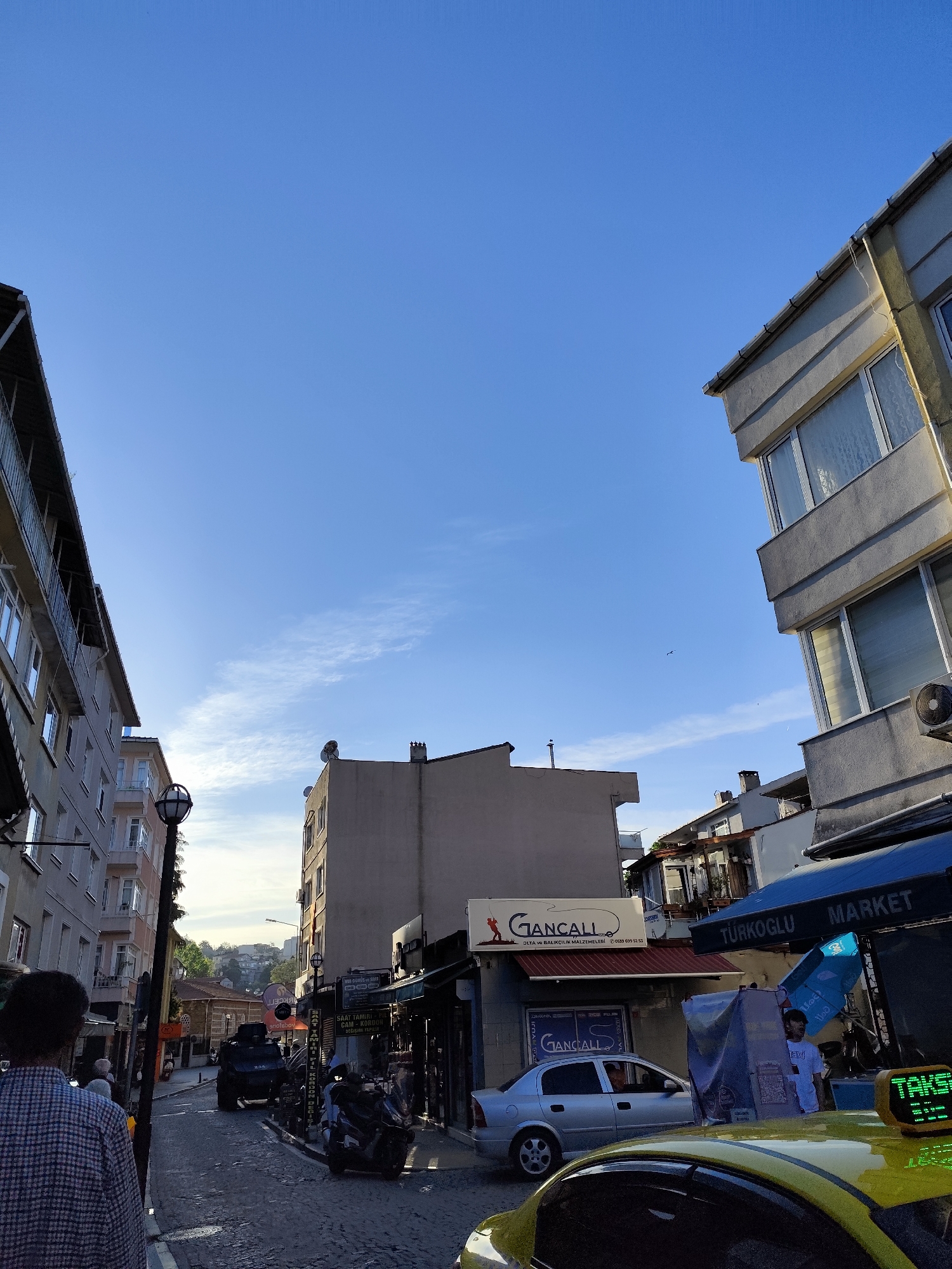 The image captures a street scene in Sarıyer, Turkey, under a clear blue sky. Buildings flank both sides of the narrow street, with a mix of architectural styles. On the right, a building with concrete-like facades and visible windows rises, partially obscured by the roof of a shop or market with a blue awning labeled "TÜRKOGLU MARKET." A man stands near the market entrance, and a yellow taxi is parked in the foreground, its "TAKSI" sign visible.
On the left, a taller building with multiple windows faces the street. The street itself is paved with cobblestones and appears to be slightly inclined. A black armored vehicle is visible further down the street, as well as a few pedestrians. A motorcycle is parked near a shop named "Gancall," identified as a fishing equipment store ("OLTA VE BALIKÇILIK MALZEMELERİ"). A silver car is parked near the Gancall shop. The image also reveals details of another shop next to Gancall that provides watch repairs, a sign of which mentions "SAAT TAMIRI," "CAM-KORDON," and "DECISIMI PAPILIR."