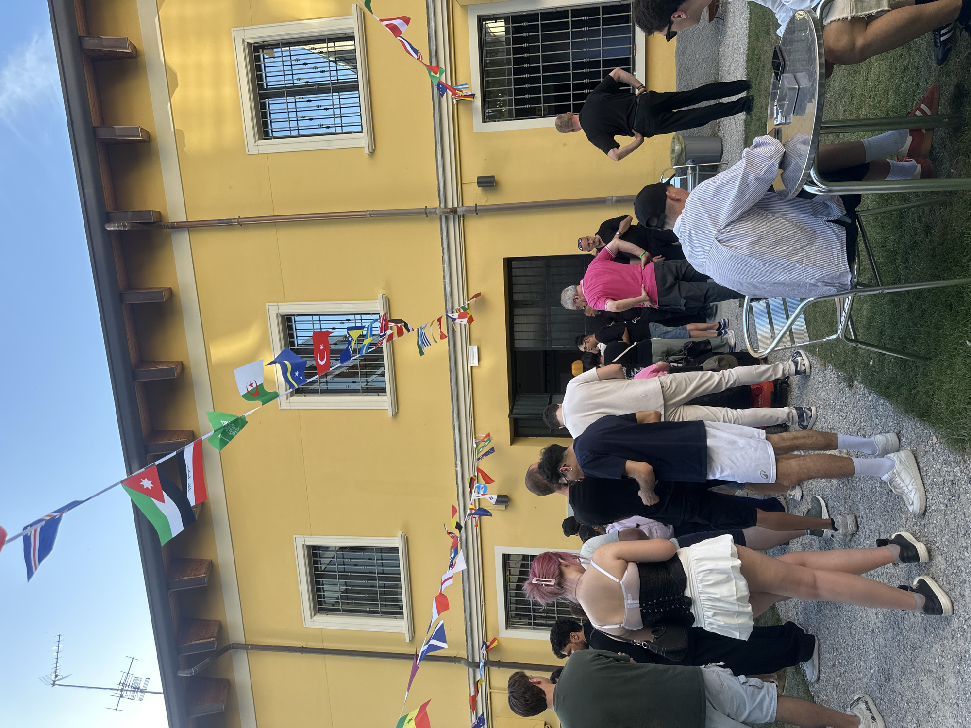 In Milan, Italy, a group of people gathers in an outdoor courtyard, likely for an event, in front of a two-story yellow building. Strings of international flags are draped across the facade and between the building and poles. The building features barred windows and wooden roof supports. In the foreground, several individuals are visible from the waist down, some standing and others seated at a round metal table with an ashtray and cell phones. The ground is covered in gravel and grass. The scene suggests a casual social gathering under a clear sky.