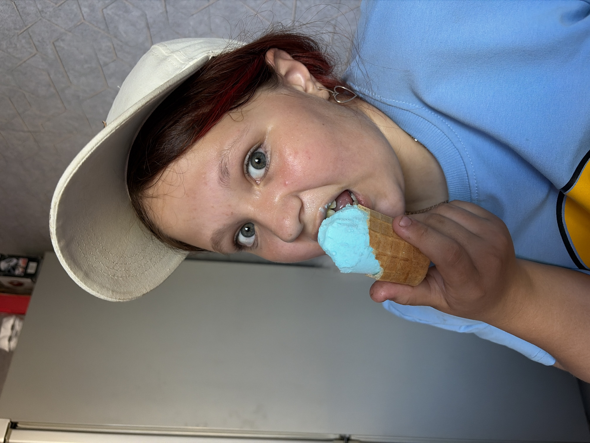 A person, seen from a tilted perspective, is eating a blue ice cream cone. The person is wearing a white baseball cap and a light blue long-sleeved shirt. They have red streaks in their hair and a silver heart-shaped earring. The background appears to be a light gray wall, potentially a refrigerator. The location is not verifiable.