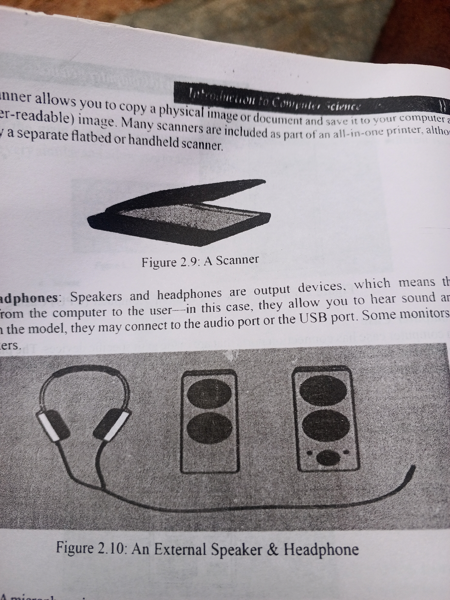 This media file is a close-up shot of a page from a book, likely a textbook on computer science. The page features printed text and two illustrations.
The top portion of the page discusses scanners, describing their function and different types. An illustration labeled "Figure 2.9: A Scanner" depicts a flatbed scanner with its lid open, showcasing the scanning bed.
The middle section of the page details speakers and headphones as output devices. Below this text is an illustration labeled "Figure 2.10: An External Speaker & Headphone". This illustration shows a pair of headphones connected by a cable to two speakers. The headphones are depicted with two earcups and a headband. The speakers are rectangular with two circular speaker elements on each.
The overall setting is a printed page, suggesting an educational context. There are no people, specific time of day, weather, or location cues visible other than the text itself. The visible text provides information about computer peripherals.
