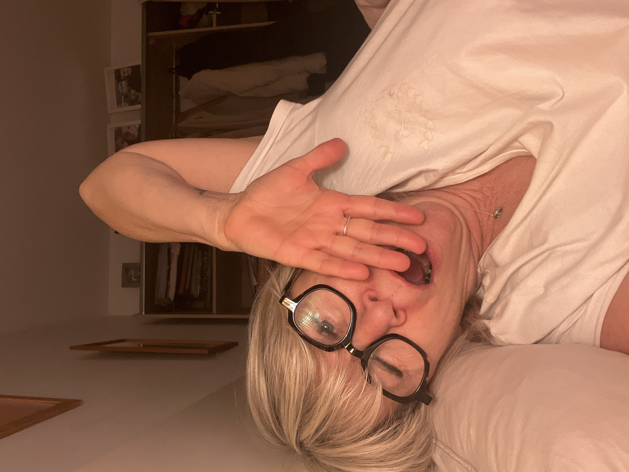 A woman with blonde hair and glasses lies on her side, facing the camera, with her right hand partially covering her face. Her mouth is open. She is wearing a white t-shirt. A silver ring is visible on her ring finger. A thin silver necklace is around her neck. In the background, there is an open closet with clothes hanging inside, as well as some framed pictures on the wall and picture frames on the surface near her. The location of Chambœuf, France cannot be determined from the image.