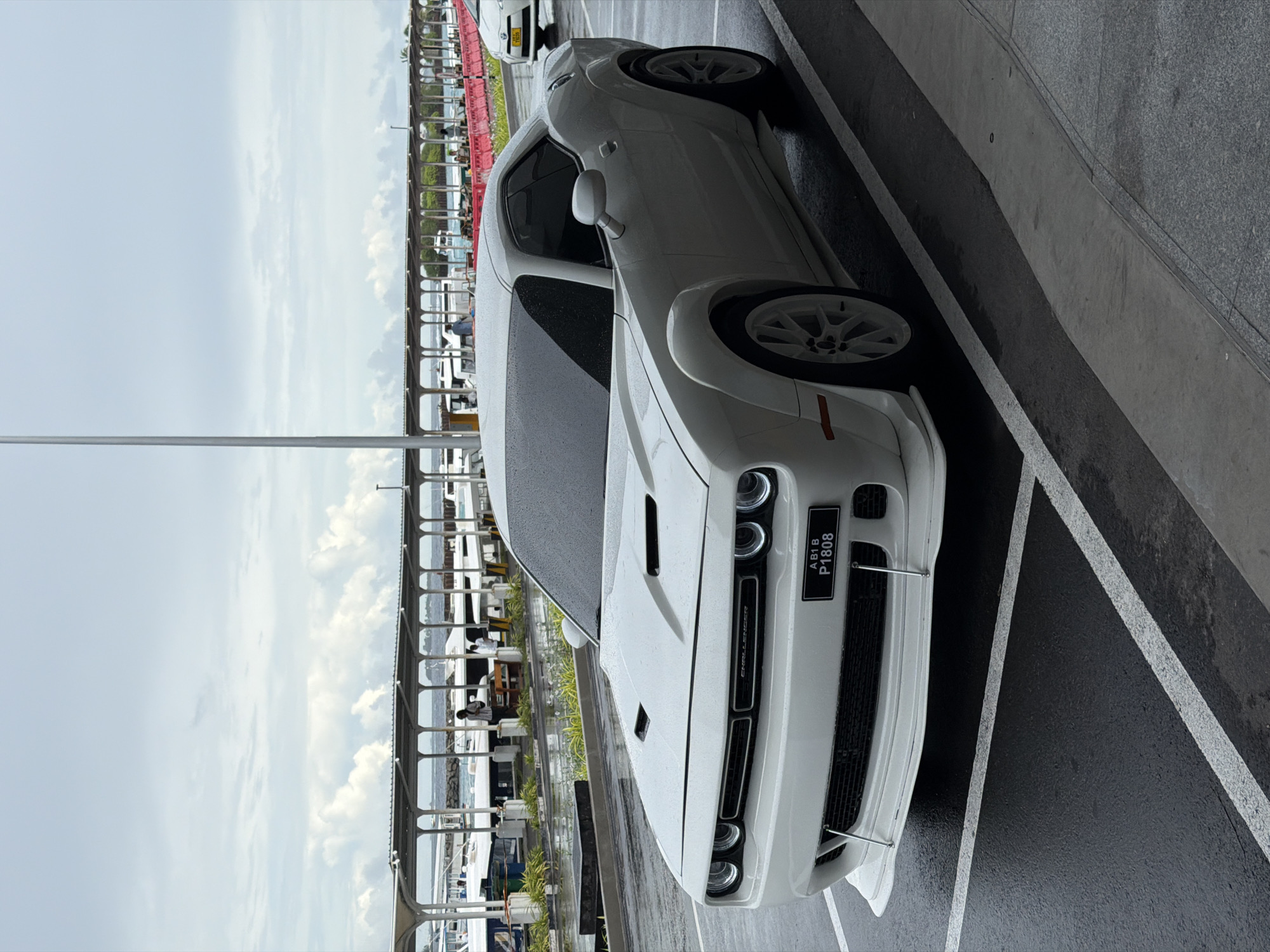 A white Dodge Challenger car is prominently featured, parked on what appears to be a wet asphalt surface, possibly after rain. The car is positioned at an angle, with its front fascia and driver's side visible. It has white multi-spoke wheels and custom wide-body fenders. On the front grille, the word "CHALLENGER" is visible. The license plate, mounted on the front bumper, displays "ABIB P1808".
In the background, a pier or railing structure runs horizontally, separating the parking area from a body of water where several boats are docked. These boats vary in size and design, suggesting a marina or harbor setting. A few people can be seen in the background near the boats and pier, some appearing to be seated or standing under a covered area. Beyond the water, a faint outline of land or buildings is visible in the distance.
The sky is partly cloudy, with a mix of light blue and white clouds, indicating daytime. The ground and car surface show moisture, suggesting recent precipitation. The scene depicts an outdoor parking lot in a coastal environment, consistent with the provided location context of Malé, Maldives.