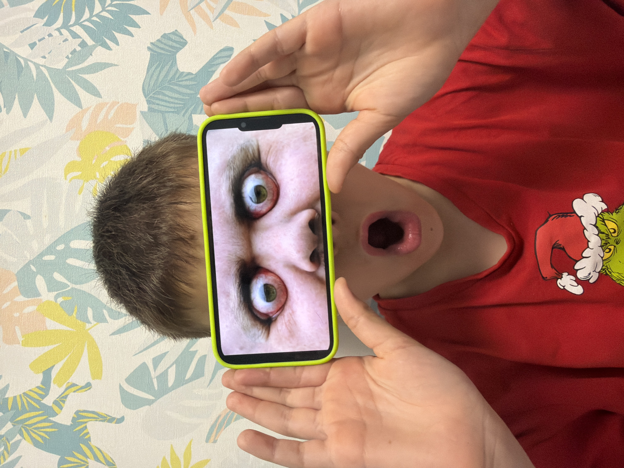 A photograph depicts a child holding a smartphone displaying a close-up image of a face with wide, bloodshot eyes and dilated pupils. The child's mouth is open, mimicking the expression on the phone screen. The child is wearing a red shirt with a graphic of the Grinch. The background features a patterned surface with floral and leaf designs. The photograph's orientation suggests it was taken from an overhead perspective. There is no information to determine the location of the image within Votkinsk, Russia.