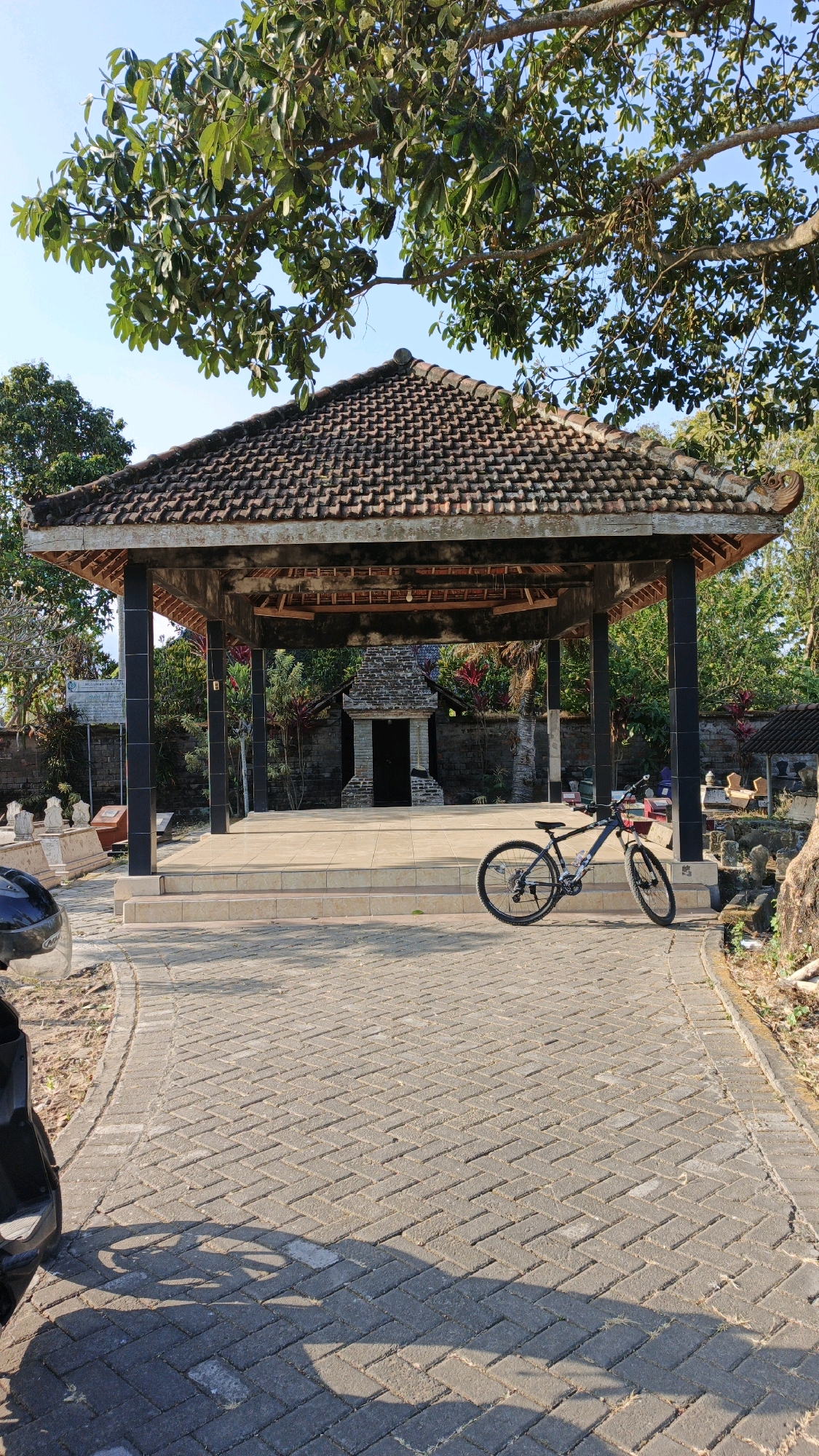 In the city of Wajak Kidul, Indonesia, a bicycle is parked on a paved walkway leading towards an open-air pavilion with a tiled roof. The pavilion has dark pillars and a raised platform. Behind the pavilion, a stone structure with a dark entrance is visible, surrounded by vegetation. The walkway is made of interlocking brick pavers, and a portion of a black car is seen on the left side of the image. Lush green trees with visible branches and leaves are overhead, with the sky visible through the foliage. The scene appears to be outdoors, under natural daylight, with shadows cast on the ground.