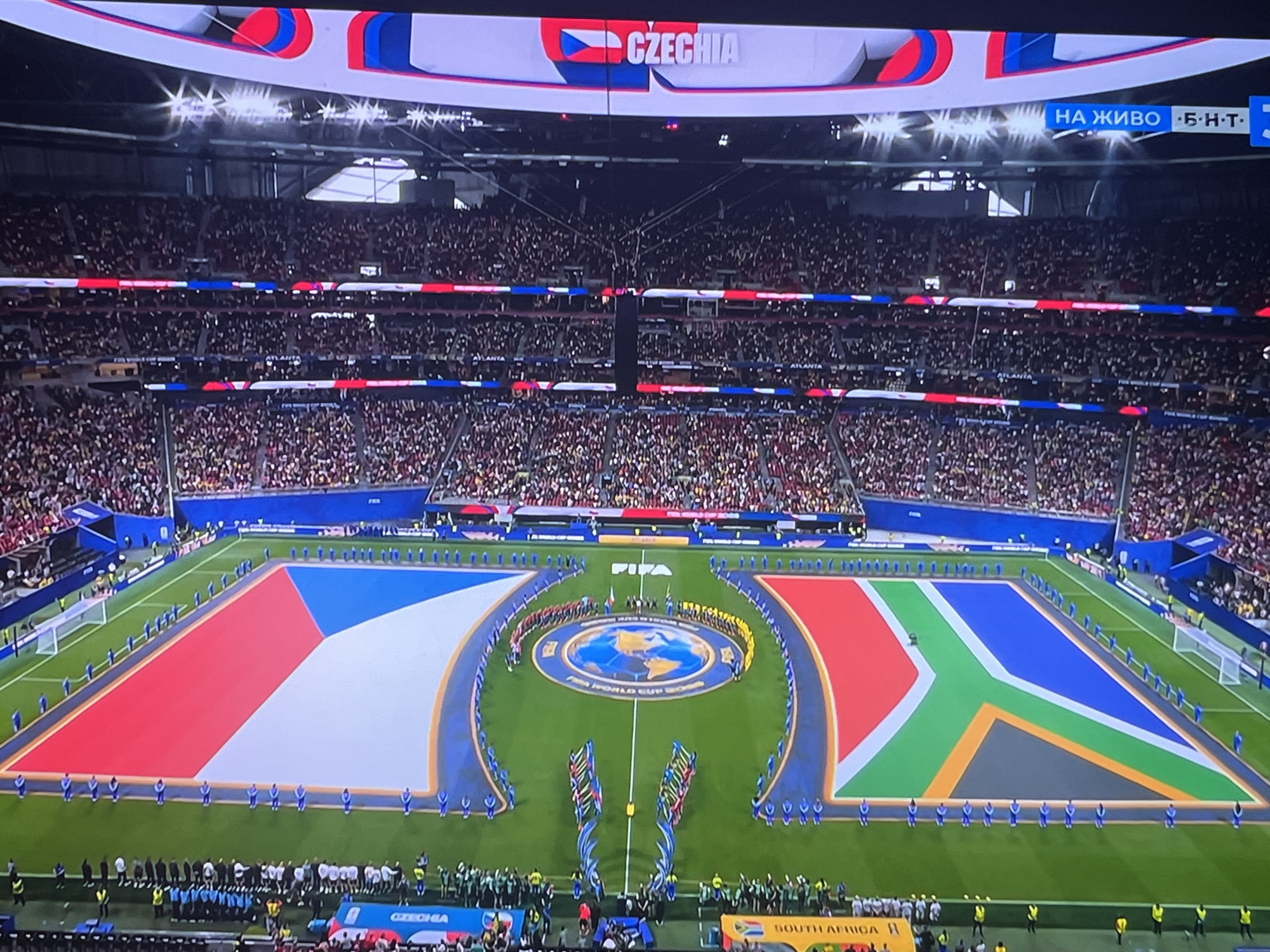 The image shows a bird's-eye view of a soccer stadium filled with spectators. The field is adorned with two large flags, one resembling the Czech flag and the other the South African flag, forming a semi-circular shape around a central circular emblem with a depiction of the Earth. Both flags are positioned on either side of a runway-like area where people are lined up. The crowd in the stadium is a mix of red and white colors, suggesting team colors or fan attire. Above the stadium, electronic billboards display text. One billboard clearly shows the word "CZECHIA" next to a Czech flag. Another billboard on the right side displays "НА ЖИВО" which is Russian for "LIVE", followed by some Cyrillic letters and a television channel logo. The atmosphere appears to be during a pre-game ceremony or a significant event. The location of Varna, Bulgaria is not discernible from the image itself.