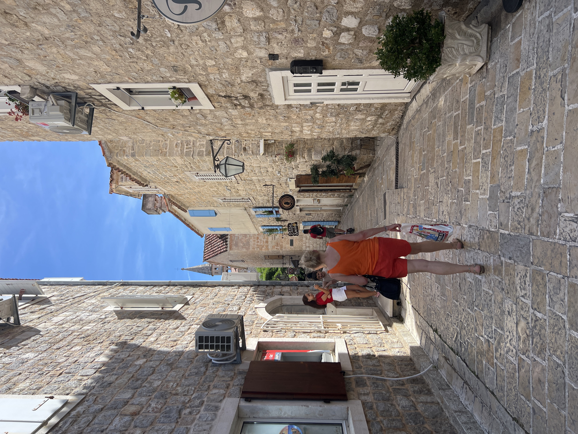 The image depicts a narrow, cobblestone street in the Old Town of Budva, Montenegro, on a sunny day. A woman in an orange tank top and red shorts walks away from the camera, carrying a shopping bag. She is walking down the street between stone buildings. To her left, a building has an air conditioning unit mounted on its exterior wall and a window with a dark brown shutter. Two other people, a woman in a red shirt and white shorts and a man, are standing near the entrance of a building to the left, in conversation. To the right, the stone wall of a building features a white door with a black mailbox. Further down the street, a church spire is visible against the blue sky. Several signs are hung on the buildings, including one for "Pizza Smiley". Plants are visible in pots and near doorways, adding greenery to the scene. The overall atmosphere is one of a charming, historic European streetscape.