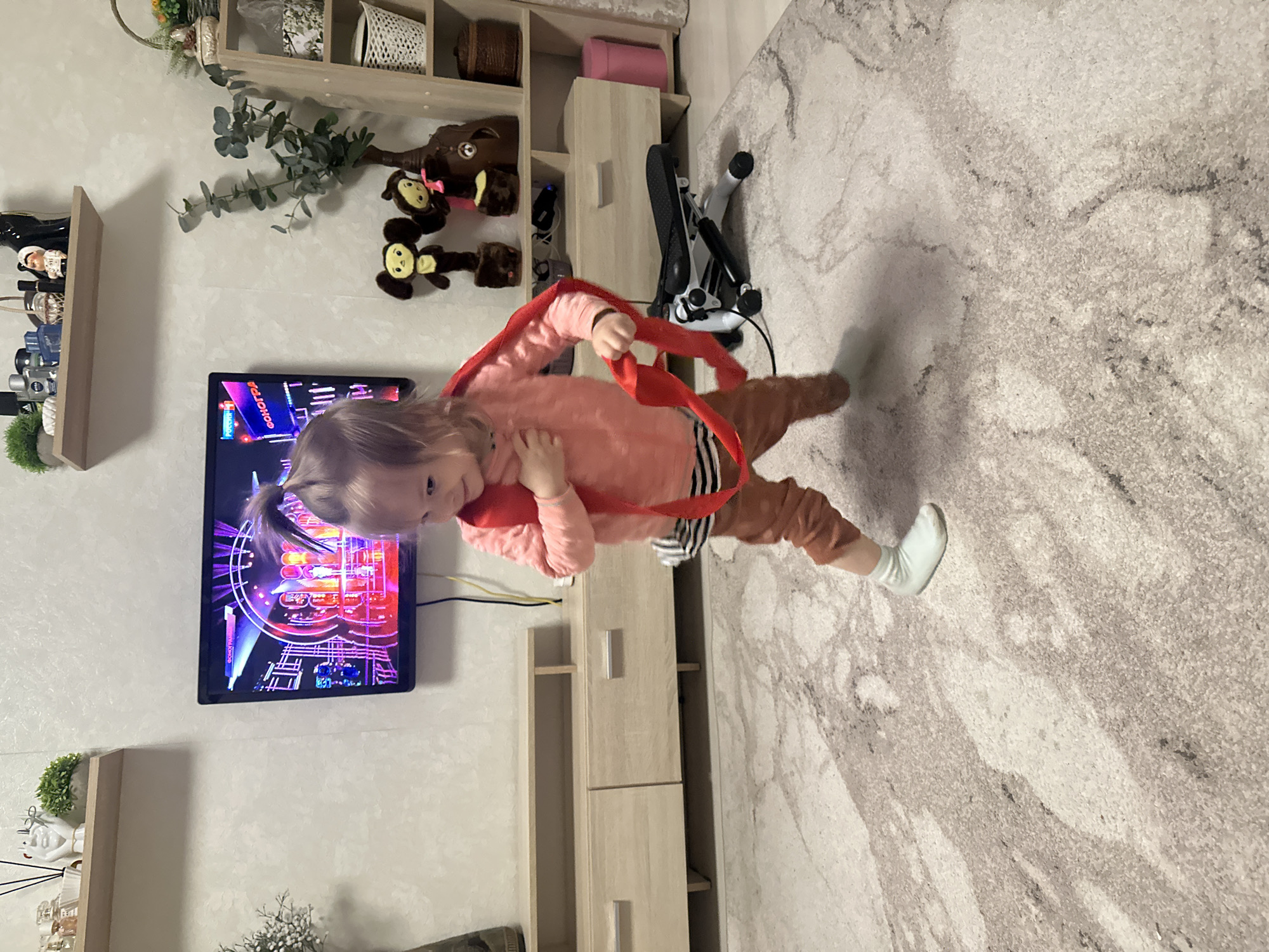 A young child with light hair tied in a ponytail stands indoors on a patterned rug. She is wearing a pink sweater, brown pants, and a white sock on her right foot. The child holds a red strap or loop in her hands, positioned around her body. A television screen on the wall behind her displays bright, colorful imagery. To the left of the TV is a small shelf with decorative items, and a larger shelving unit is on the right. A piece of exercise equipment is visible near the shelving unit. The location appears to be a home in an urban area, likely Uva, Russia.