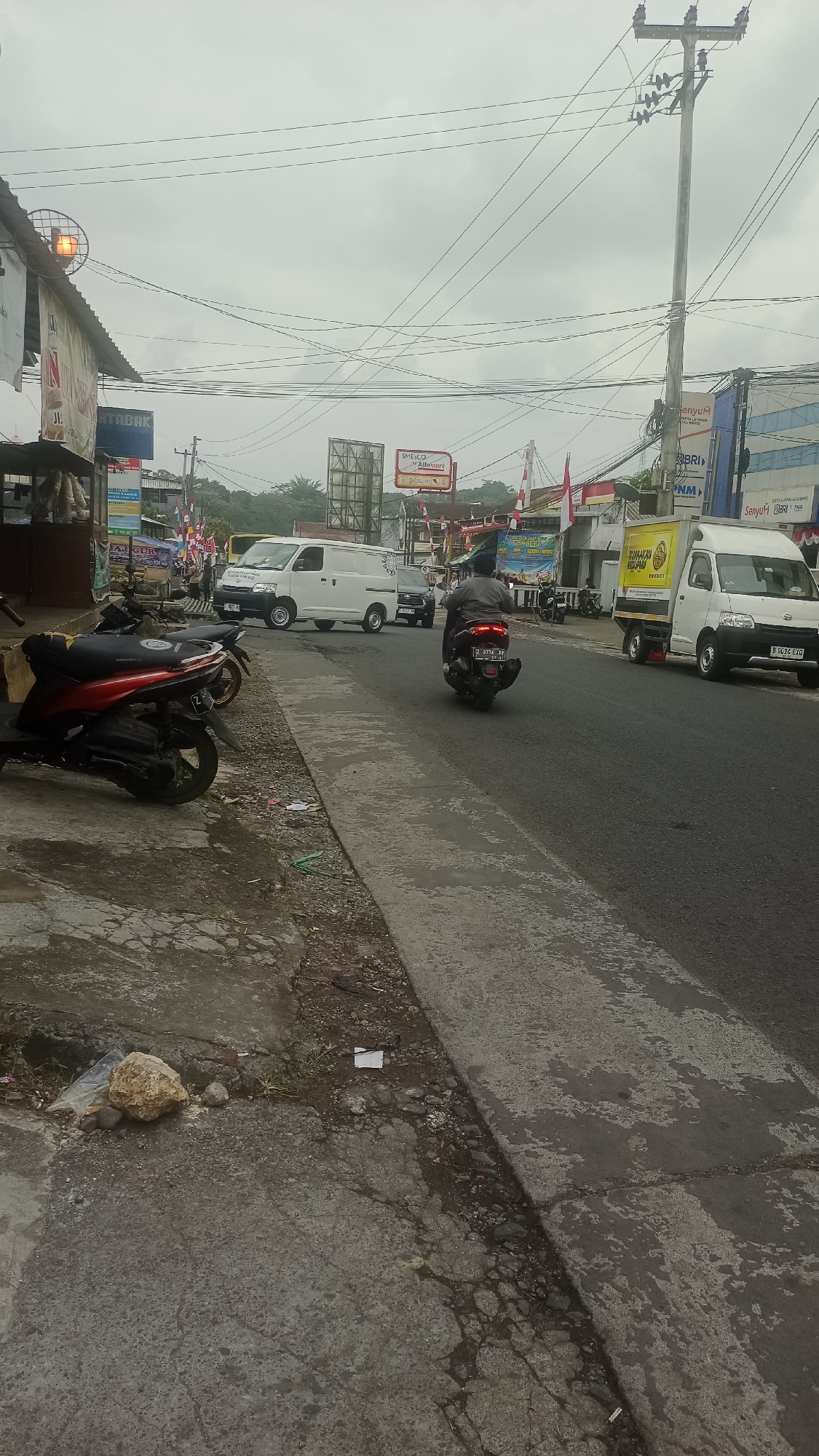 The image captures a street scene in Karyamandala, Indonesia, under an overcast sky. In the foreground, two parked motorcycles are visible on a paved sidewalk adjacent to a road. One motorcycle is red and black, and the other is black. The sidewalk appears weathered and cracked, with scattered debris and a large rock.
The road itself features several vehicles. A black motorcycle with a rider wearing a dark jacket and helmet is moving away from the viewer. Further down the road, a white van is traveling in the same direction, followed by another white van. A white truck with a yellow cargo box is parked on the right side of the road.
Several buildings and signs line the street. On the left, a building with a sign that reads "RTABAK" is partially visible. There are also various banners and flags, including Indonesian flags. On the right side of the road, buildings with signs for "Alfamart" and "BRI" are apparent. Overhead, a dense network of electrical wires crisscrosses the sky, supported by utility poles. The overall environment suggests a commercial or semi-urban area.