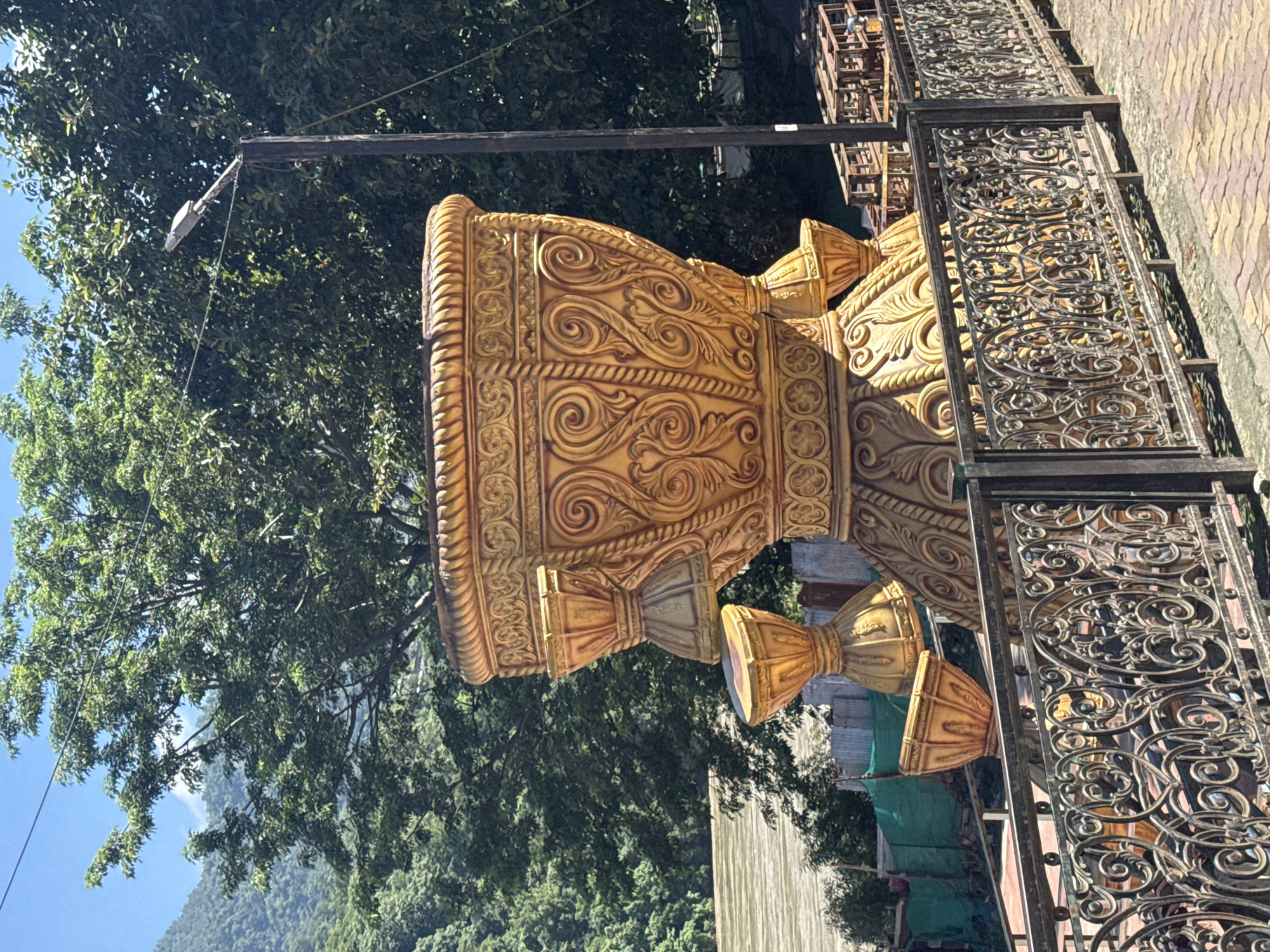 A large, ornate golden statue of a pot with decorative patterns and multiple tiered sections stands prominently on a paved walkway in Rishikesh, India. The statue features intricate swirling designs and a rope-like border. Several smaller, bell-shaped golden elements are attached to the main structure, some appearing to cascade downwards. The statue is positioned behind a decorative black metal fence with elaborate scrollwork. In the background, lush green trees and a mountainous landscape are visible under a clear blue sky. A body of water, possibly a river, flows in the middle ground. A street lamp is visible on a dark pole to the right of the statue. Construction materials, including wooden pallets, are stacked behind the fence in the middle ground. The paving stones in the foreground are a mix of red and yellow hues. No people are visible in the image.