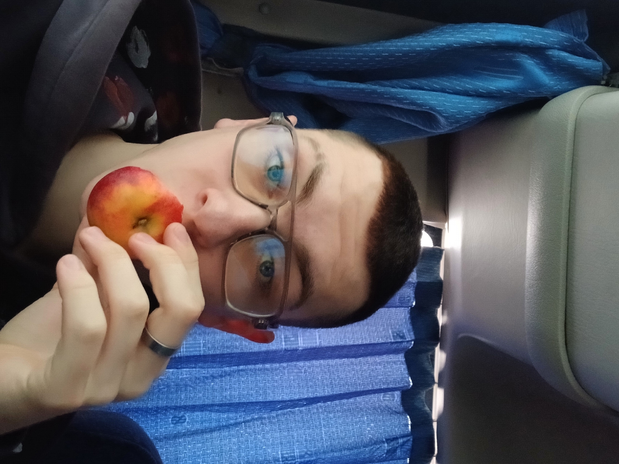 A person with short dark hair and glasses holds a peach near their face. They are wearing a ring on their left hand. The background includes blue fabric, a grey headrest, and window blinds. There is no evidence to confirm the image was taken in the city of Gory, Russia.