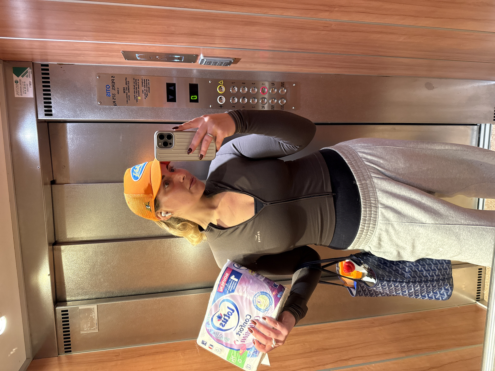 A woman takes a mirror selfie in an elevator with wooden walls and a stainless steel door, possibly in Paris, France. The elevator's control panel shows buttons for floor levels 0 through 5, with level 1 illuminated. The woman wears an orange baseball cap, a gray zip-up athletic shirt, and gray sweatpants. She holds a smartphone with a tan-colored case in her left hand and a package of "Autof Comfort" toilet paper in her right. A navy blue tote bag is slung over her shoulder.