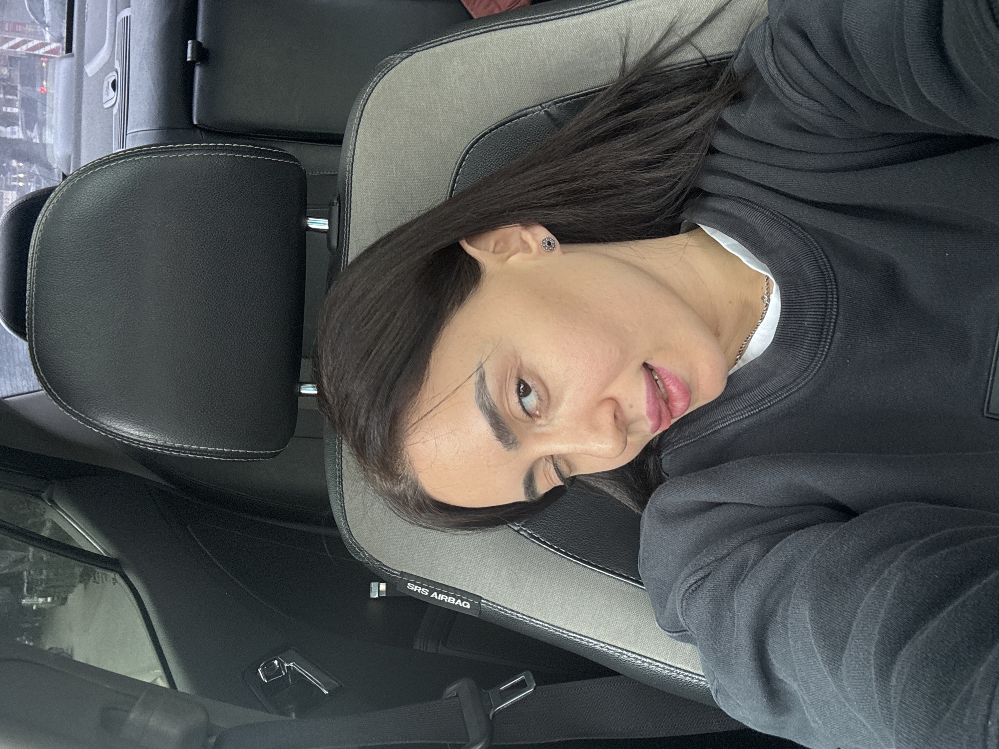 The image depicts an adult female positioned in the front passenger seat of a vehicle, located within the city of Tbilisi, Georgia. The subject, with dark, shoulder-length hair, is wearing a dark crewneck sweatshirt over a white undershirt, a silver chain necklace, and a circular stud earring in her right ear. She is looking directly at the camera, winking with her right eye, and has her tongue slightly extended. The vehicle's interior features a two-tone passenger seat with black leather and grey fabric, displaying an "SRS AIRBAG" label. A black seatbelt is visible on the left. The background visible through the car windows shows a blurry urban street environment with other vehicles and structures, indicative of a daytime scene.