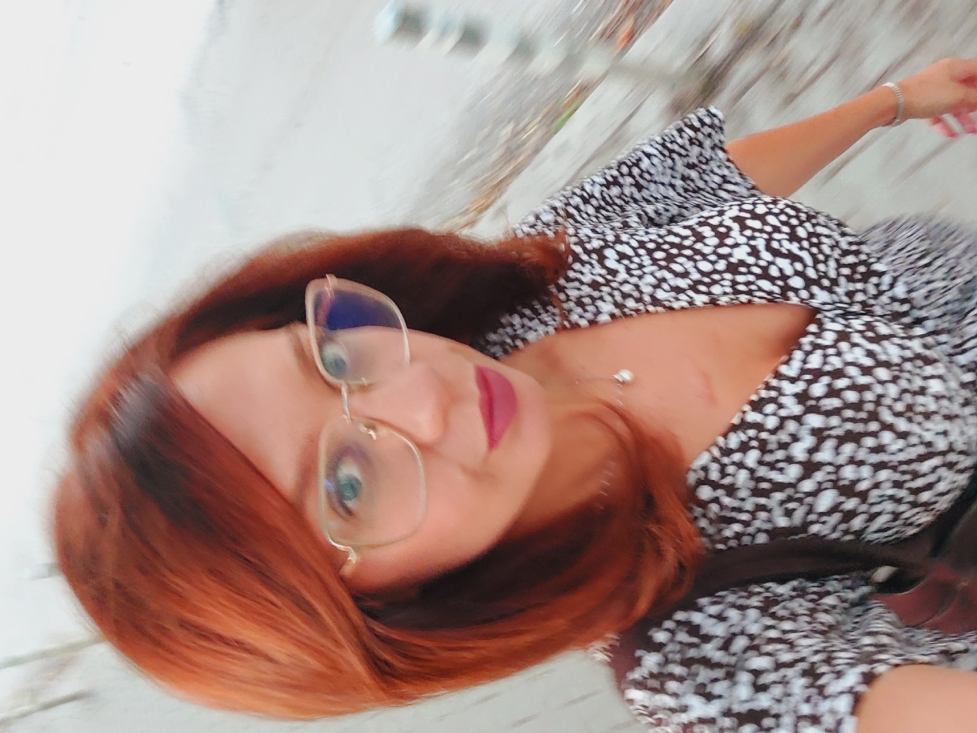 A woman with shoulder-length auburn hair and blue eyes is taking a selfie outdoors. She is wearing glasses with thin gold frames, a necklace with a small pendant, and a dark-colored dress with a white speckled pattern. A part of a brown bag is visible. The background is blurred and appears to be a walkway or path with railings on the right side. The overall lighting suggests it is daytime, with overcast conditions. There are no discernible landmarks or indicators to identify the specific location within Sofia, Bulgaria.