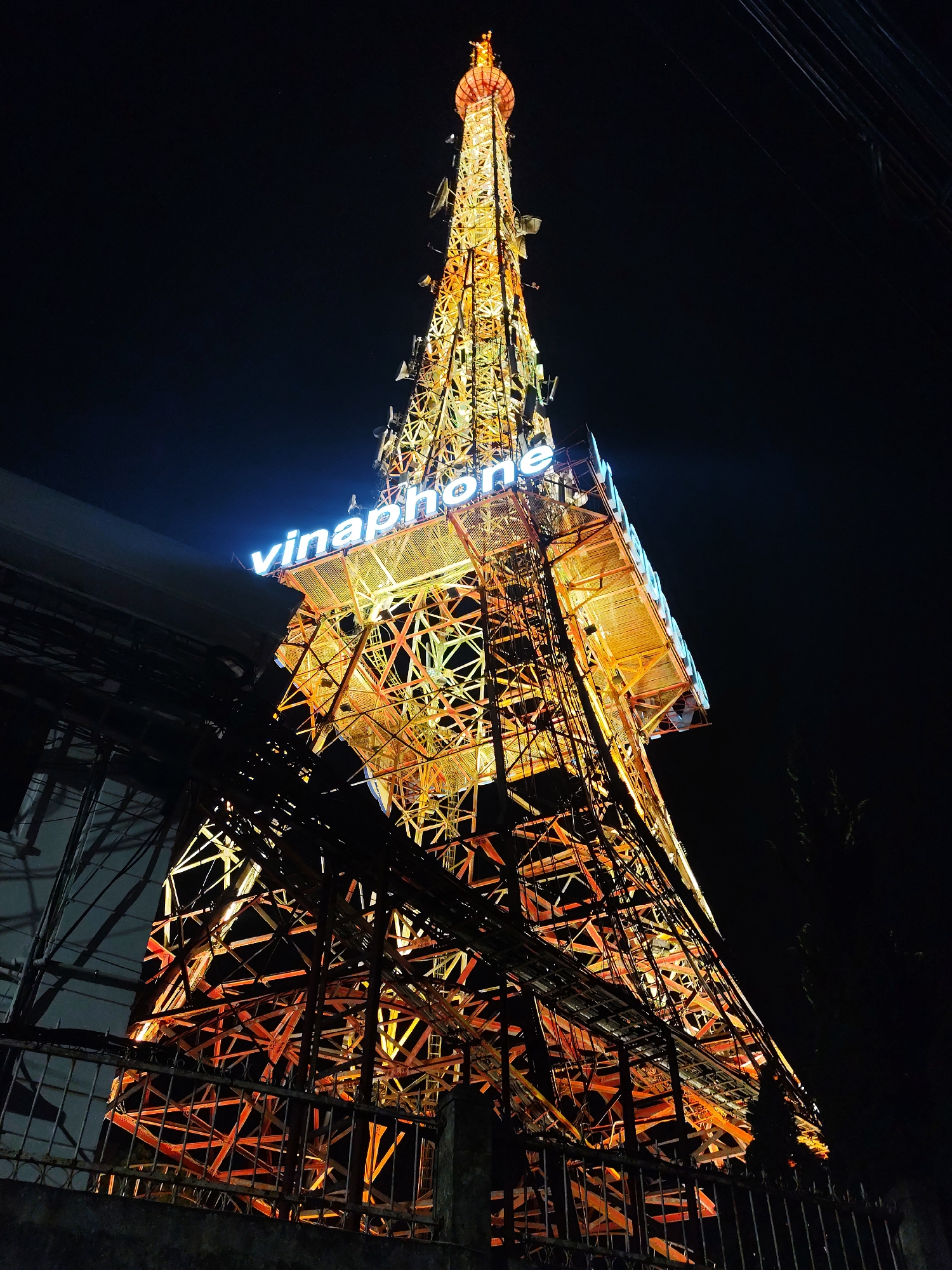 This image captures the Vinaphone Tower in Da Lat, Vietnam, at night. The tower, a prominent landmark, is illuminated with bright orange and yellow lights, highlighting its intricate lattice structure. A neon blue sign displaying "vinaphone" is visible near the top of the tower. The surrounding environment is dark, indicating nighttime, with some electrical wires and a fence visible in the foreground. The perspective is looking up at the tower from a low angle. There are no people or discernible actions depicted in the scene.