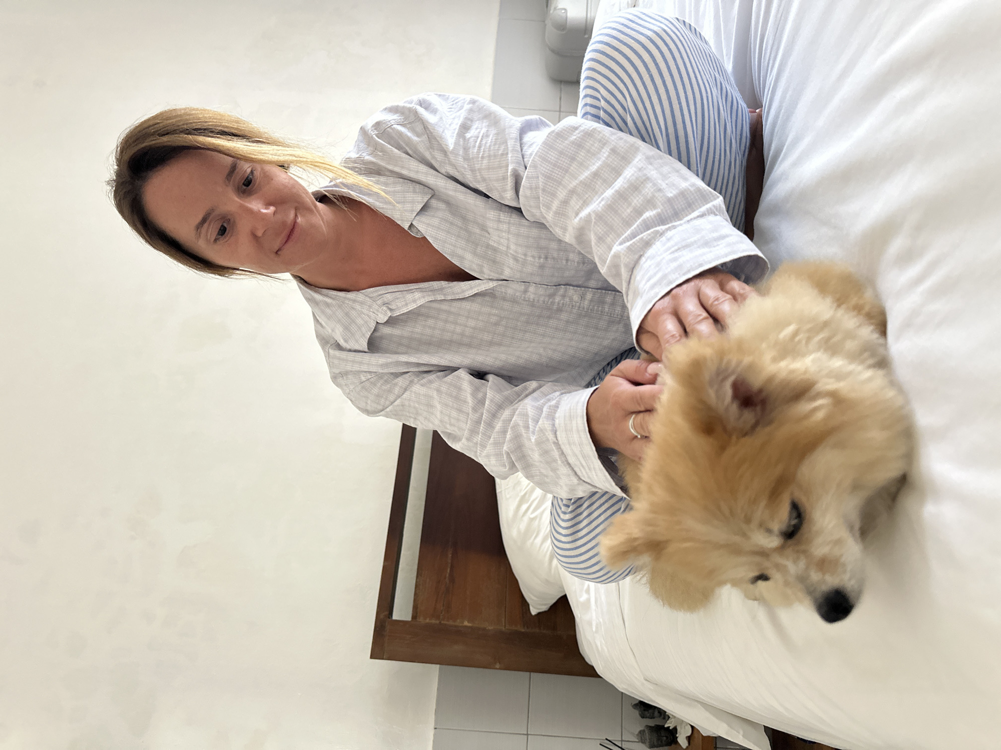 In a bedroom in Kuta Selatan, Indonesia, a woman sits cross-legged on a white-sheeted bed with her Pomeranian dog. The woman is wearing a light blue and white plaid pajama set and has her hands on the dog. The dog is light brown and white, with its face turned to the left. The background features a wooden headboard, a white wall, and a partially visible bedside table.