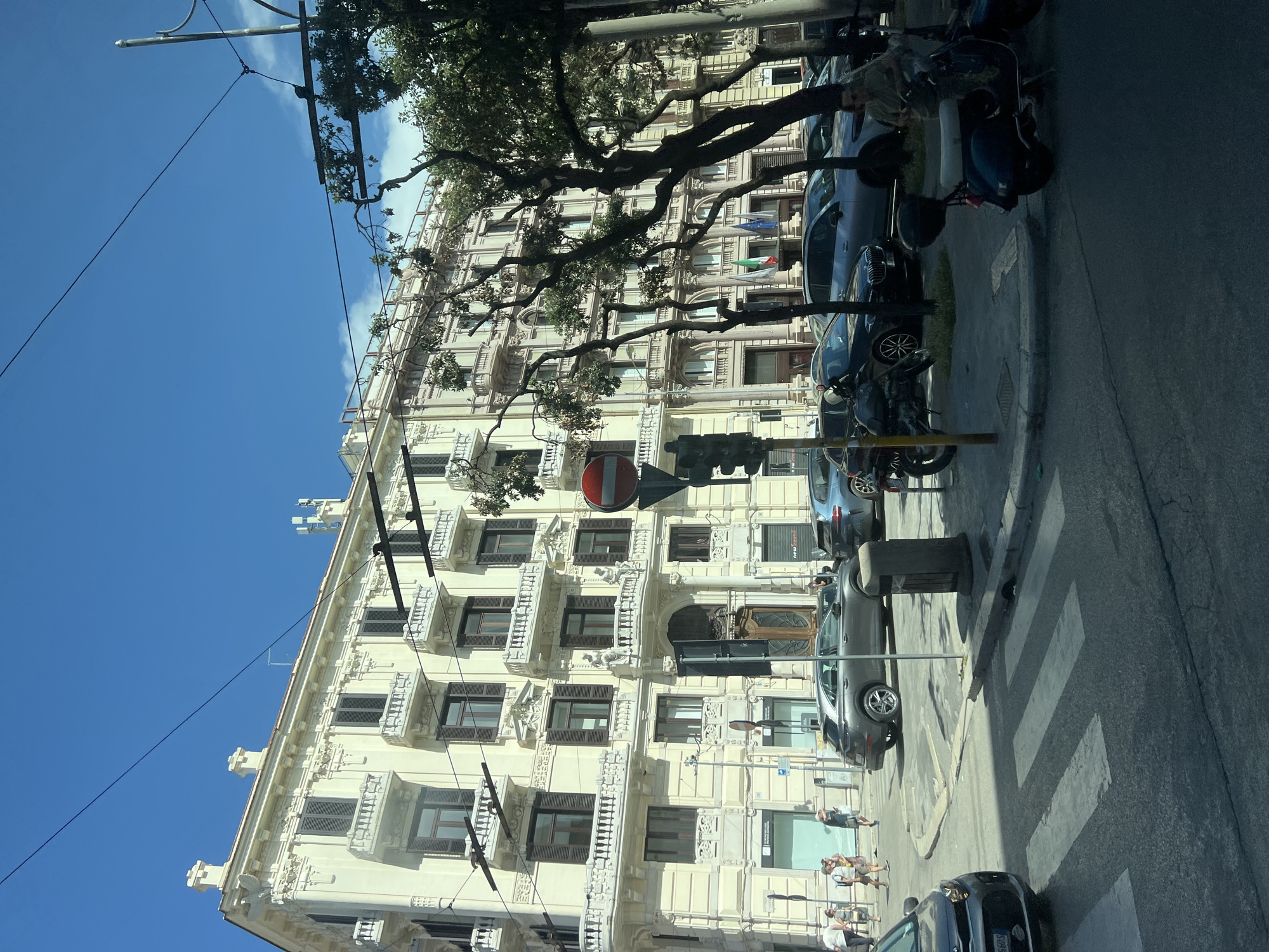The image depicts a street scene in Bari, Italy. A large, ornate, cream-colored building with multiple balconies and dark shutters dominates the background. In the foreground, a street with a crosswalk is visible. Several cars, including a dark gray SUV and a dark hatchback, are parked or driving on the street. Two motorcycles, one dark and one a dark blue scooter, are also present. A traffic light and a circular red and white sign are on a pole to the right of the crosswalk. Several pedestrians are walking on the sidewalk near the building. Trees with dark green foliage are scattered throughout the scene. The sky is clear and blue with some wispy clouds.